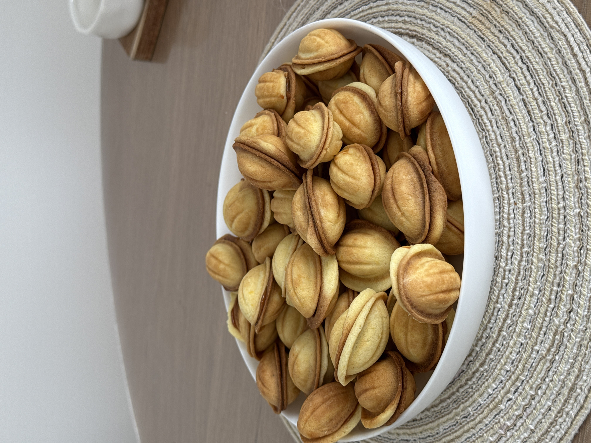 A high-angle shot captures a white bowl overflowing with numerous golden-brown, walnut-shaped cookies. These cookies, likely "Oreshki," are composed of two halves joined together, with visible seams and a slightly darker filling peeking through on some. The bowl rests on a light-colored, circular woven placemat, which has a distinct textured pattern of concentric braids or rows.
In the background, a warm-toned wooden surface (possibly a table or shelf) is partially visible to the left of the bowl, extending towards the upper left corner. Adjacent to this wooden surface, a plain white wall or counter creates a minimalist backdrop. In the far upper-left corner, a small portion of a white cup sits on a small wooden coaster or shelf, adding a subtle domestic detail. The lighting is soft and even, suggesting an indoor setting. There are no people or discernible activities, and no visible text in the image. The overall scene conveys a sense of quiet readiness, as if the cookies are prepared for serving.