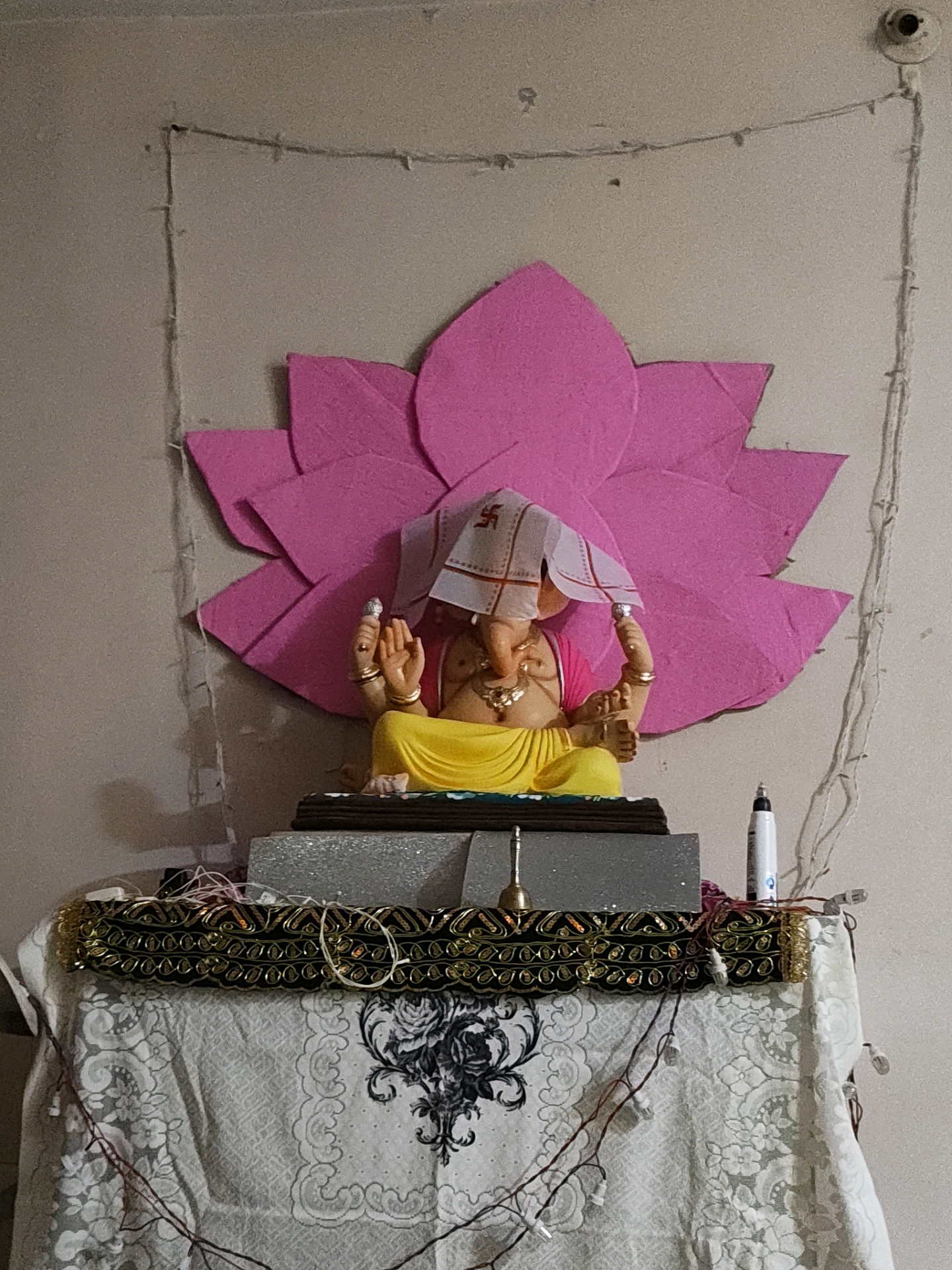 The image shows a statue of the Hindu deity Ganesha placed on a decorated altar. The statue is adorned with a yellow garment and a white cloth draped over its head, which features a swastika symbol. Behind the statue, a large pink lotus flower decoration is mounted on a light-colored wall. The altar is covered with several decorative cloths, including a black and gold patterned fabric above a white lace-like cloth with floral designs. Various small bells, wires, and string lights are also visible around the altar and extending onto the wall. A white marker pen stands upright on the right side of the altar. The background appears to be a plain, light-colored wall. The overall scene suggests a domestic setting, possibly a home altar for religious worship. The specific location in Pune, India cannot be determined from the image.