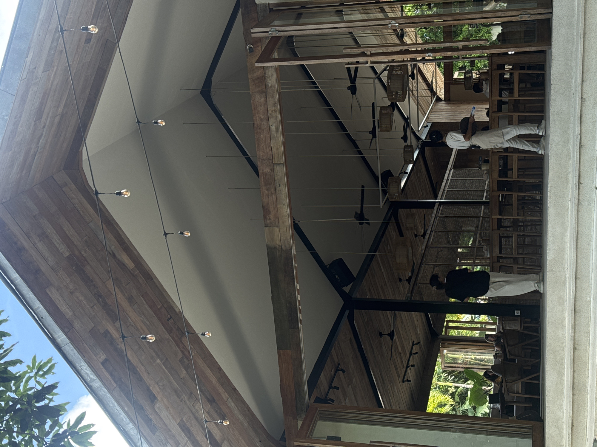 Captured from a low-angle perspective, the image showcases the interior of a building in Kuta Selatan, Indonesia. The architectural design emphasizes a high, angular wooden ceiling with exposed light fixtures, creating a contrast with the white walls beneath. Dominating the right side of the frame, a large window provides a glimpse into a restaurant or seating area, with wooden furniture and patrons visible. Two individuals are distinctly visible: one dressed in white clothing and the other in black. These figures are seen seated at tables, suggesting a relaxed dining environment. The presence of lush greenery outside the window offers a tropical aesthetic, reinforcing the location's ambiance in Indonesia.
