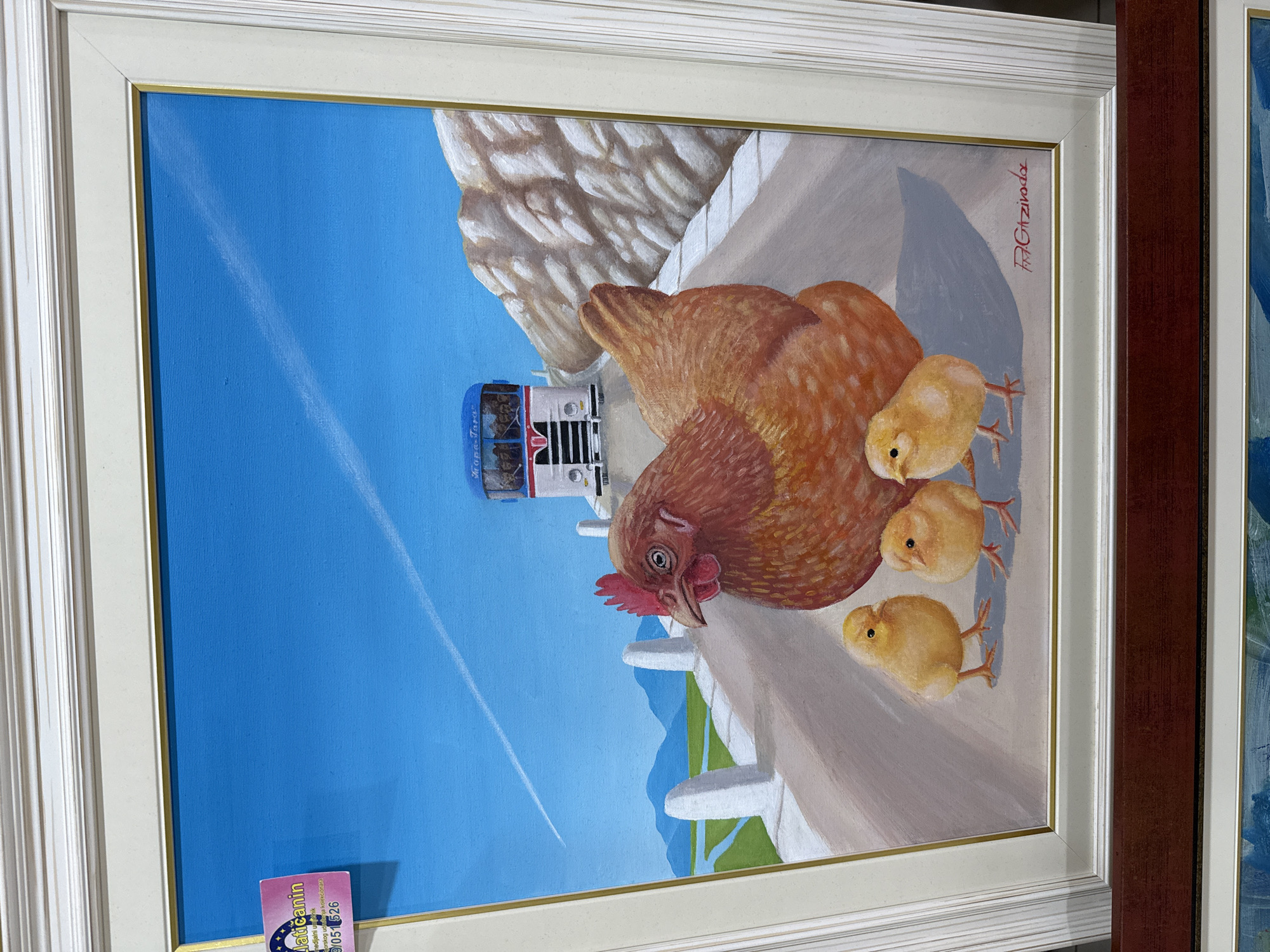 The image shows a framed oil painting of a hen and three chicks on a road, with a bus and mountains in the background. The hen is in the foreground, facing left with its head turned towards the viewer. The chicks are gathered around its feet. The road appears to be in a rural setting, with white posts lining its edge and green fields and mountains in the distance under a bright blue sky with a contrail. A white bus with "Tops-Tours" written on its side is seen on the road behind the hen and chicks. The painting is signed in the lower right corner.