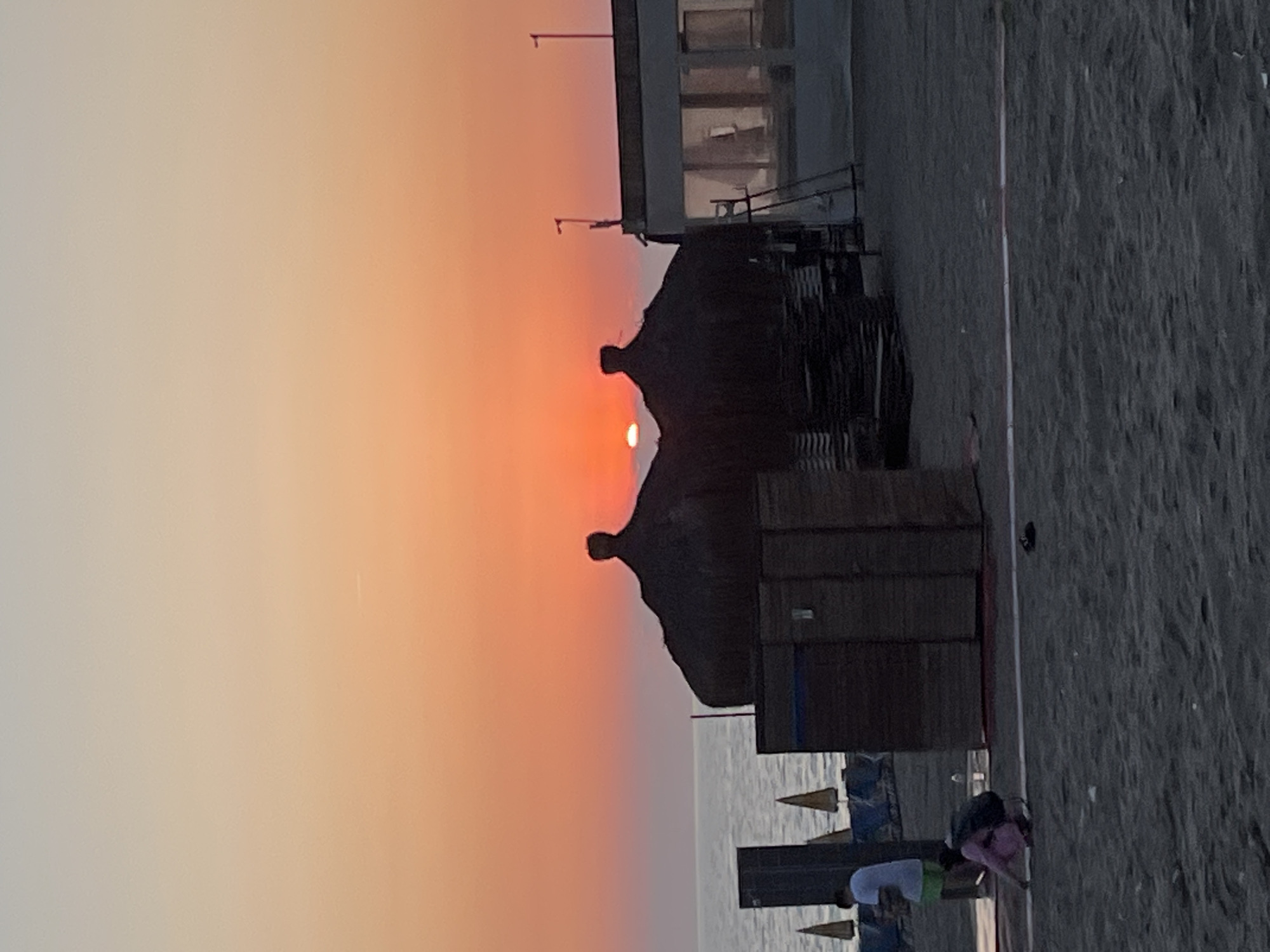 Here is a description of the image:
The image appears to be taken at a beach in Alanya, Turkey, featuring a sunset over the Mediterranean Sea. The sky has a gradient of color from a pale orange to a darker orange. The sun is partially obscured by thatched beach umbrellas or huts, silhouetting them against the sky. There is a building slightly out of focus in the background. Closer to the foreground, there is a beach with people. The people include someone wearing white and green clothing and someone wearing a pink top.