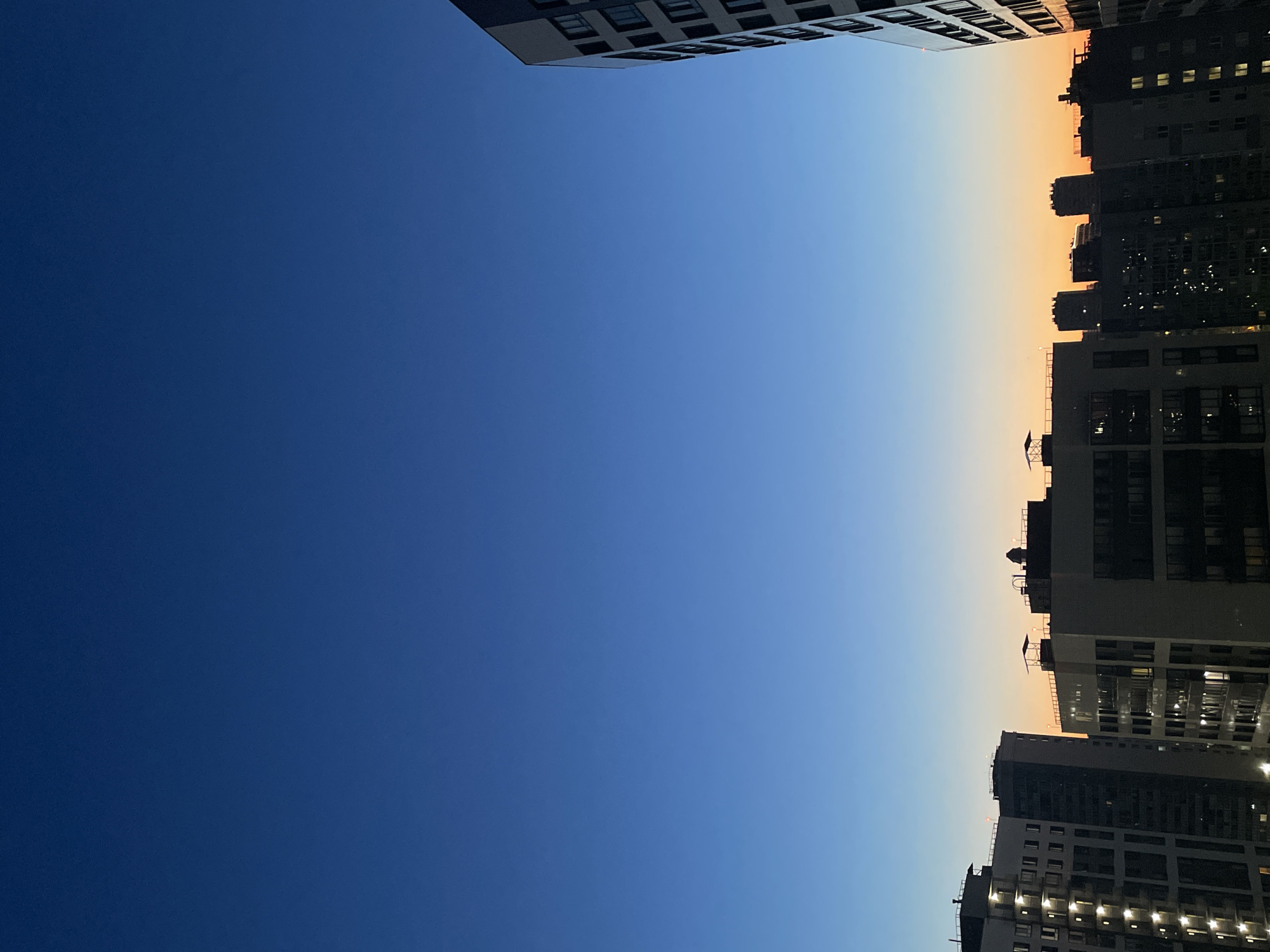 The image shows a cityscape at dusk, likely in Sinyavino, Russia, given the context. The sky transitions from a deep blue at the top to a faint orange hue near the horizon. Silhouetted against the twilight are several apartment buildings of varying heights and architectural styles. Some buildings have their windows illuminated, suggesting residents are home. The overall impression is one of urban life settling down for the night.