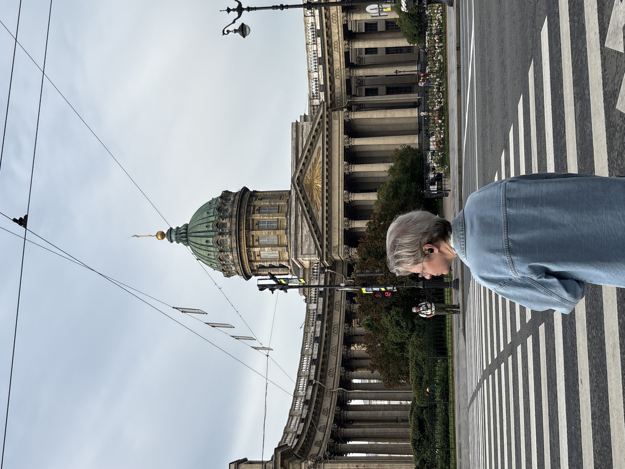 A person with grey hair and glasses stands on a pedestrian crossing, facing away from the camera. They are wearing a light blue denim jacket. Behind them, a grand building with a green dome and classical architecture is visible. The building features a long colonnade and ornate details. The sky is overcast. There are traffic lights and street lamps in the vicinity. Several other people and some greenery are present in the background. This scene is likely Saint Petersburg, Russia, specifically in front of Kazan Cathedral, identifiable by its distinctive dome and semi-circular colonnade.