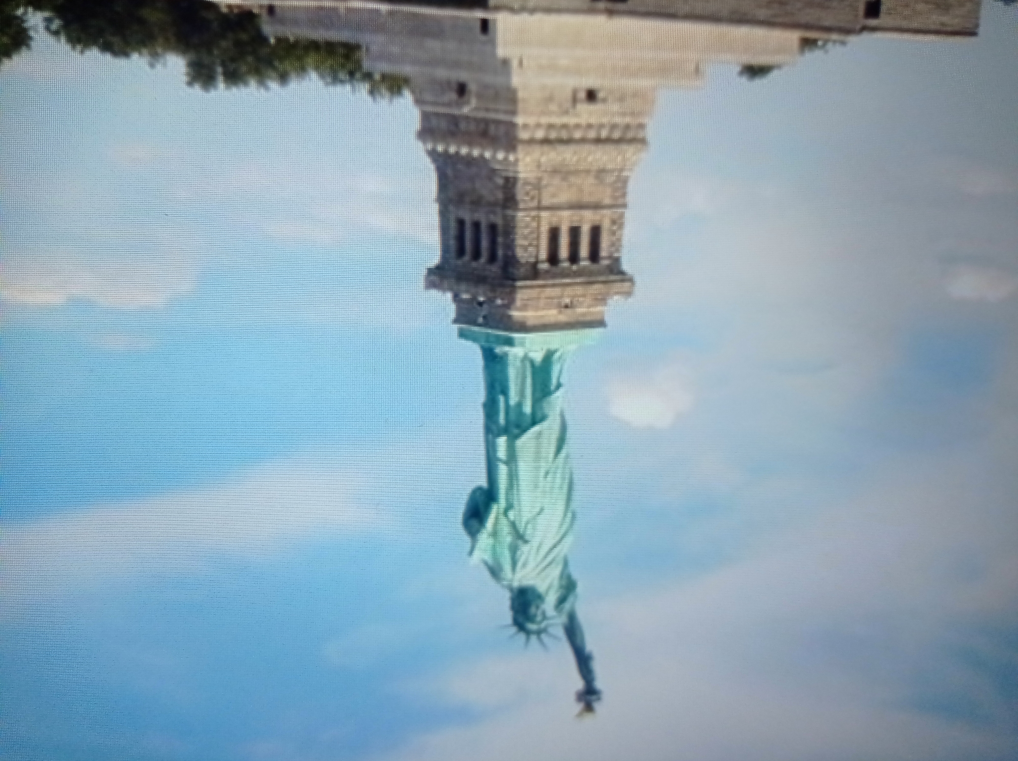 The image depicts the Statue of Liberty against a bright blue sky with scattered clouds. The statue, made of a greenish oxidized copper, stands tall on a stone pedestal. The pedestal is intricately designed with rectangular openings and decorative elements. In the foreground, partially visible at the bottom, are parts of a stone structure, possibly representing a base or an island, with some greenery and trees peeking out. The lighting suggests it is daytime. The scene is a clear, static shot of the iconic monument. There is no indication of the city of Mukachevo, Ukraine in this image.