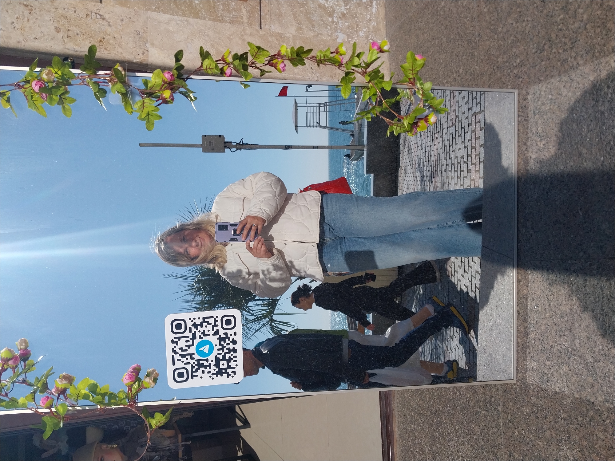 A mirror selfie is captured outdoors, showing a woman with blonde hair, wearing a white quilted jacket and blue jeans, holding a purple smartphone to take the picture. She stands in front of a large, rectangular mirror decorated with a green garland of artificial pink flower buds and leaves draped across the top and bottom.
The mirror reflects a bright, sunny coastal scene. In the background, a blue sky meets the sparkling blue sea. A white lifeguard stand with a red flag is visible on the beach. A paved walkway or promenade runs alongside the water. Also reflected in the mirror are two other individuals walking: one person in dark clothing with short dark hair, and another person in a dark jacket and blue jeans with distinct yellow shoes. The ground where the mirror is placed appears to be a textured, grey-brown stone.
A prominent detail is a white square QR code with the blue and white Telegram logo in its center, positioned in the lower-left quadrant of the mirror's reflection. The time of day is clearly sunny, indicated by the bright sky and reflections. The overall impression is a vibrant outdoor setting on a clear day. The image itself is rotated 90 degrees clockwise, with the woman and reflections appearing on their sides.