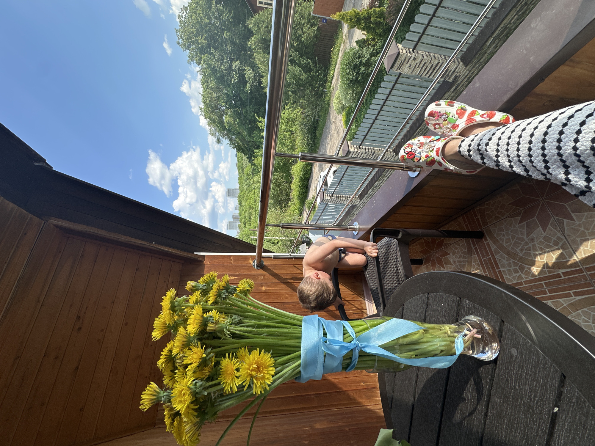 A sunny outdoor scene shows a child peering over the edge of a balcony railing. A large bouquet of dandelions in a glass vase sits on a dark-colored patio table beside the child. The dandelions are tied with a light blue ribbon. The balcony has a metal railing and a wooden and metal fence overlooking trees and buildings. The background landscape appears to be within the city of Nizhny Novgorod, Russia, as tall buildings are visible in the distance. A person wearing strawberry-patterned clogs and black and white patterned pants is also on the balcony. The balcony floor has a tiled pattern. The side of a wooden structure is visible, providing shelter to the balcony.