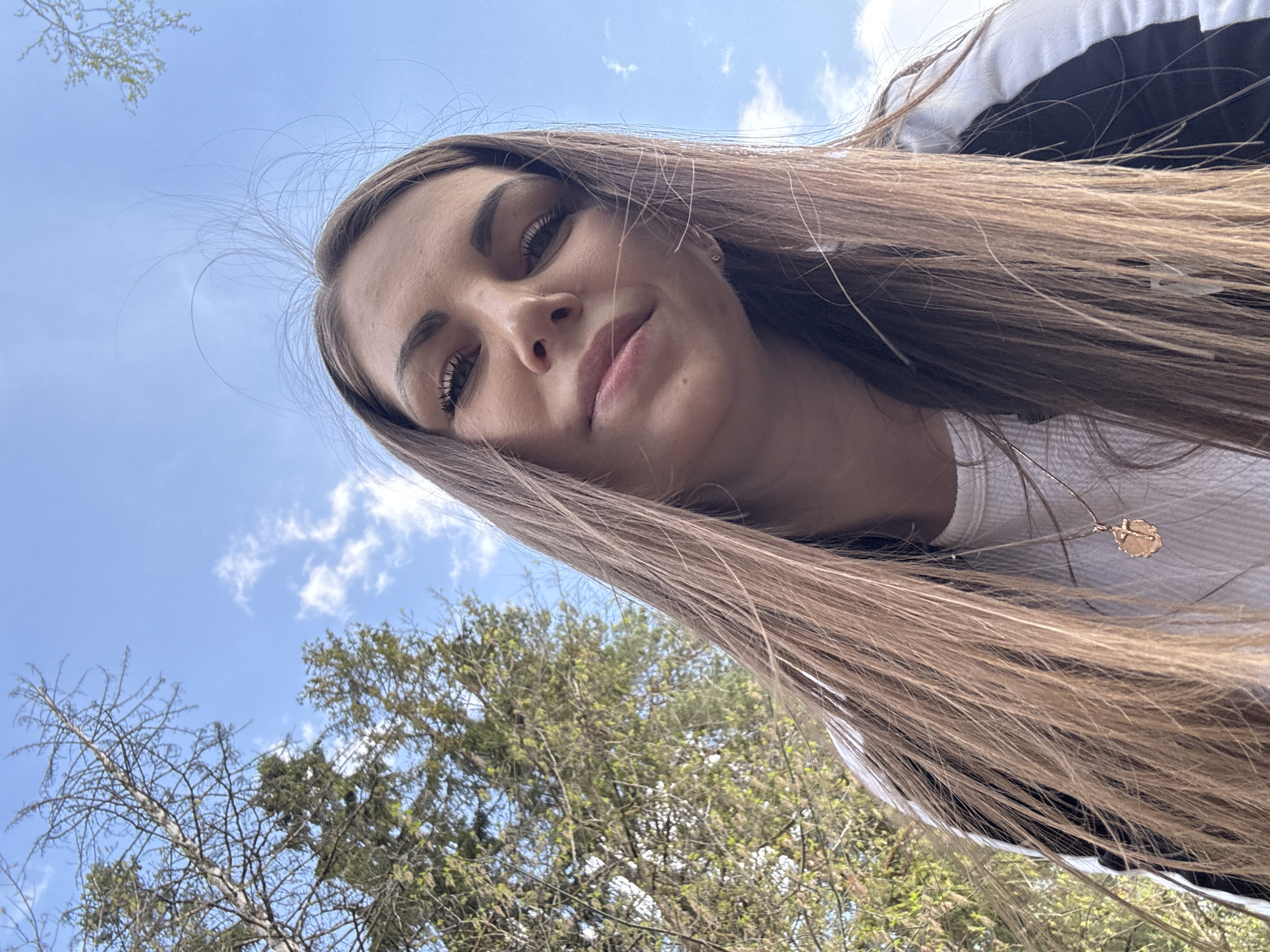 A low-angle shot shows a woman with long, brown hair gazing downwards towards the camera. She is wearing a white, ribbed shirt and a black jacket with white trim. A gold necklace with a round pendant is visible around her neck. Behind her, there is a clear blue sky with scattered white clouds, as well as tree branches with some leaves budding out. The location is implied to be Razdolye, Russia, though no specific landmarks or identifying details are present in the image. The woman's face is the primary subject of the photo.