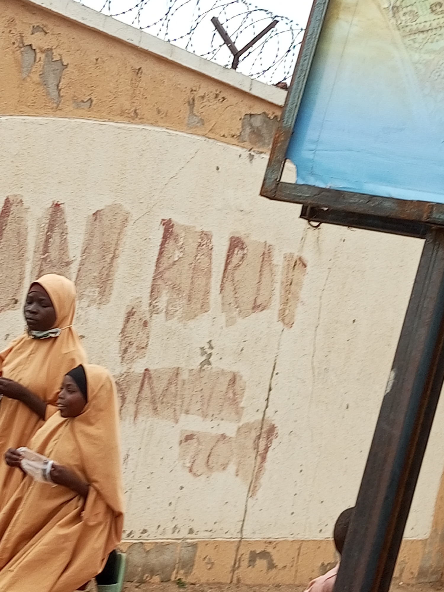 In Pantame, Nigeria, two women are seen outdoors. The woman in the foreground is wearing a light orange hijab and robe, and she is holding a clear plastic bag. Behind her, a second woman, also in a light orange hijab and robe, is looking to the left. They are standing in front of a textured wall with peeling paint, which is topped with barbed wire. To the right of the frame, a portion of a blue sign on a metal stand is visible.
