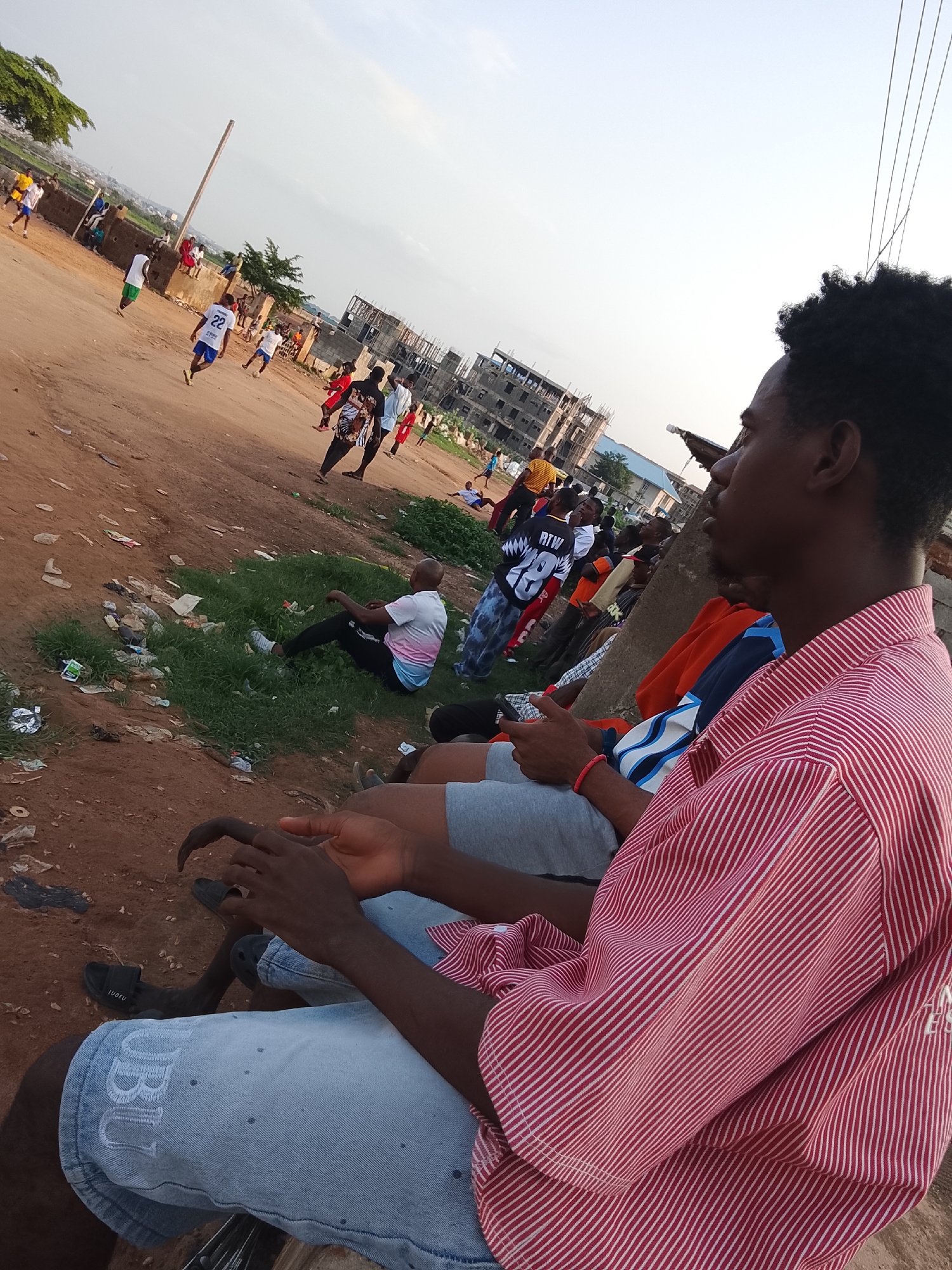 The image depicts an outdoor scene in Abuja, Nigeria, where a group of young men are gathered. In the foreground, a man wearing a red and white striped shirt and light blue denim shorts is seated, holding a smartphone. To his left, another person is visible from the waist down, wearing grey shorts and resting their hands. Behind them, several other individuals are seated and standing.
In the middle ground, a group of young men are engaged in a game of soccer on a dirt field. Some are actively playing, while others appear to be observing. The field is uneven and marked by patches of grass and scattered debris, including plastic waste. Further in the background, there are buildings under construction, indicating ongoing development in the area. The sky is overcast, suggesting it might be late afternoon or early evening. The overall environment appears to be a developing urban or semi-urban area.