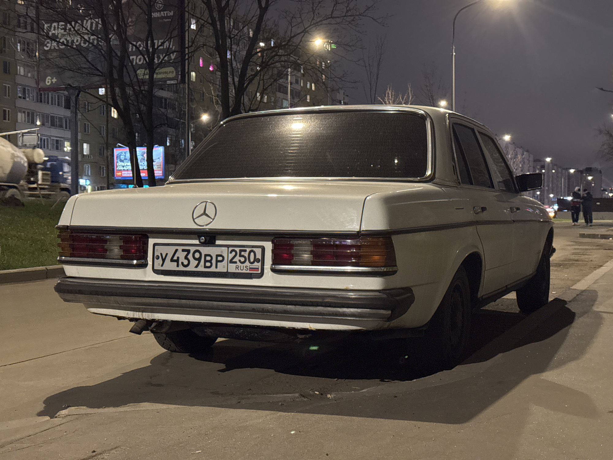 The image depicts a white Mercedes-Benz W123 parked on a street at night. The car's rear is prominently displayed, showcasing its taillights, bumper, and license plate, which reads "У439ВР 250 RUS."
In the background, a city scene unfolds with multi-story buildings, bare trees, and streetlights illuminating the area. A concrete mixer truck is visible on the left, while two pedestrians are seen walking in the distance on the right. Billboards displaying Russian text are also present.
Based on the license plate and architectural style, the location is likely Moscow, Russia. The overall atmosphere is urban and dimly lit, suggesting a quiet nighttime scene.