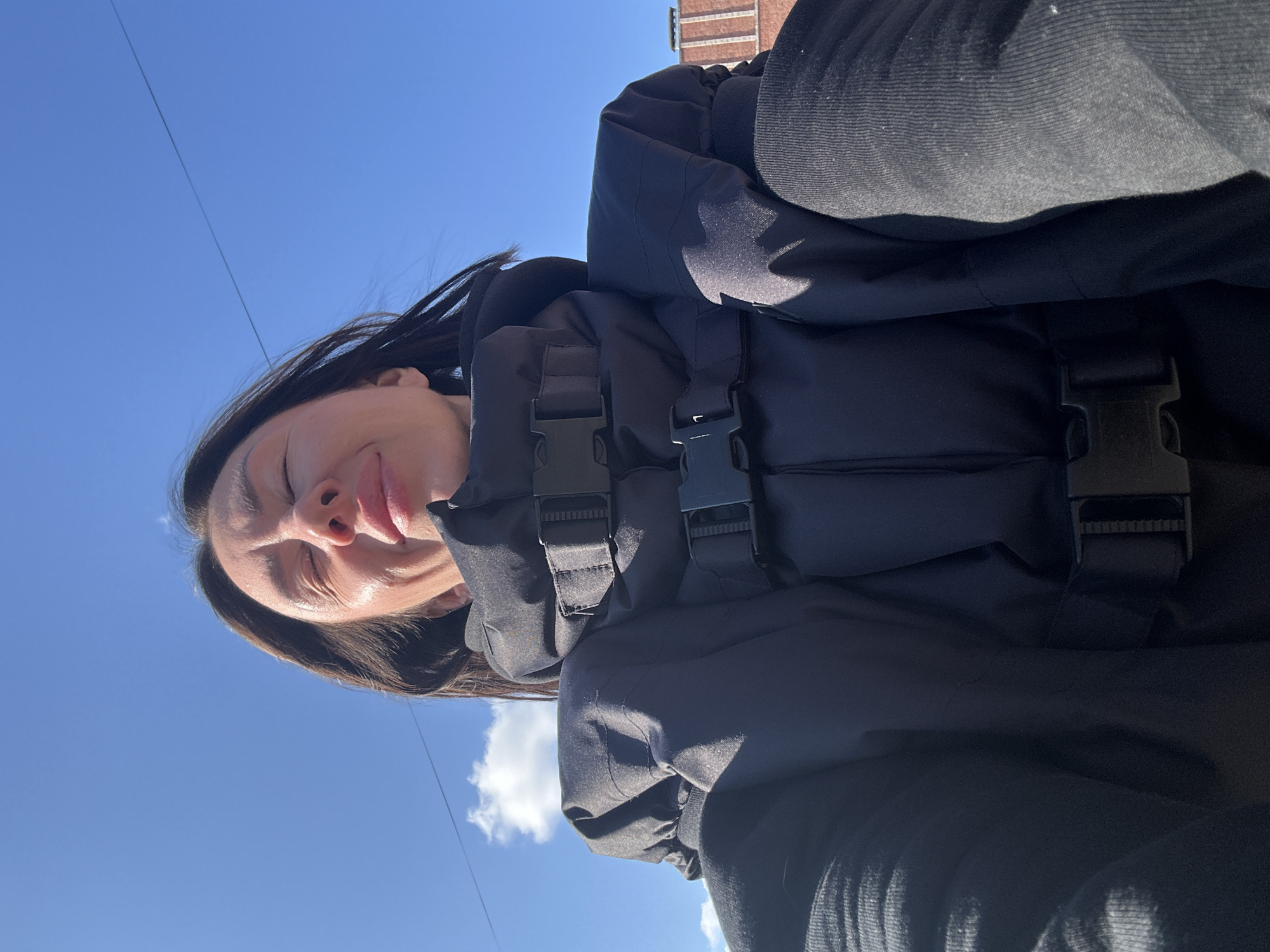 A low-angle, tilted outdoor shot features a woman in a black jacket with buckle detailing. She appears to be squinting, possibly due to sunlight. The background consists of a clear blue sky with a few clouds and a building visible in the upper right corner of the frame. There is no information to specify the exact location within Votkinsk, Russia.