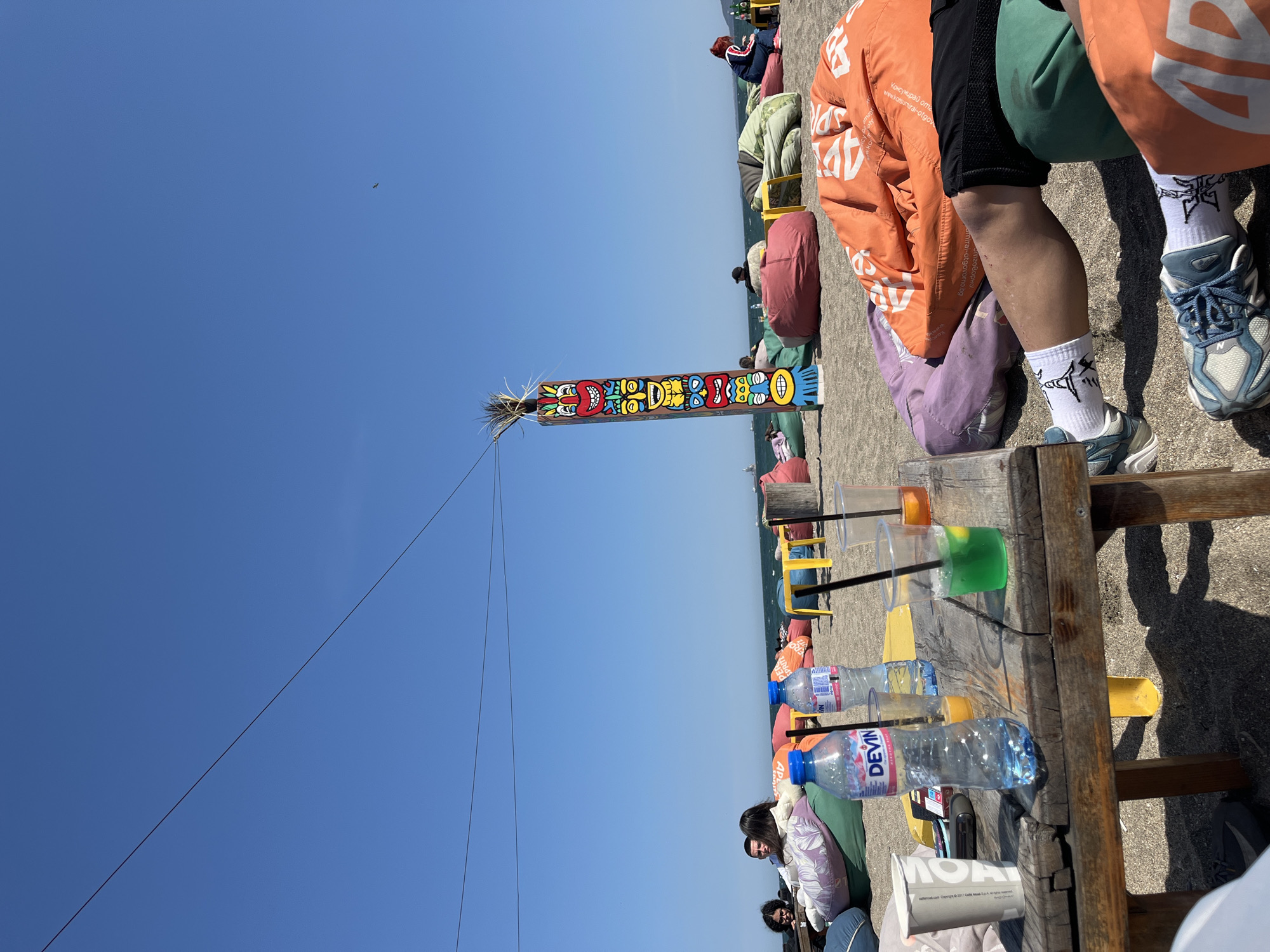 This image captures a vibrant, sunny outdoor scene at a beachside cafe or lounge in Burgas, Bulgaria. The setting is bright and clear, indicating midday with strong sunlight casting shadows.
In the foreground, a wooden table holds several items: two plastic bottles of "DEVIN" mineral water, two plastic cups with colorful beverages (one orange, one bright green, both with straws), and a white paper cup with "MOA" printed on it. The table surface is worn wood.
The perspective is from a low angle, looking towards a colorful, tall totem pole adorned with cartoonish faces and stylized letters, including "D" and "O". The totem pole stands prominently, adding a playful element to the environment.
Beyond the table and totem pole, several people are visible, relaxing on large, colorful bean bags and low chairs on what appears to be sand or light gravel. The bean bags are in shades of orange, purple, green, and red, some featuring Cyrillic text, such as "КОНЦУМИРАЙ ОТГОВОРНО" and a website address "www.kamenitza.bg" on an orange jacket and bean bag, likely advertising a Bulgarian beer brand.
A person's legs and feet are prominent in the right foreground. They are wearing black shorts, white socks with a distinctive black graphic design, and light blue and white athletic sneakers (New Balance brand is visible on one shoe). An orange jacket is draped over their lap/legs.
The background reveals the vast, clear blue sky, with a small bird soaring high above. Thin black lines or wires stretch across the sky, possibly part of a canopy or lighting structure. The horizon line shows a faint body of water (the sea) in the distance, confirming the coastal location. The overall atmosphere is relaxed and leisurely, suggesting people enjoying a sunny day outdoors.