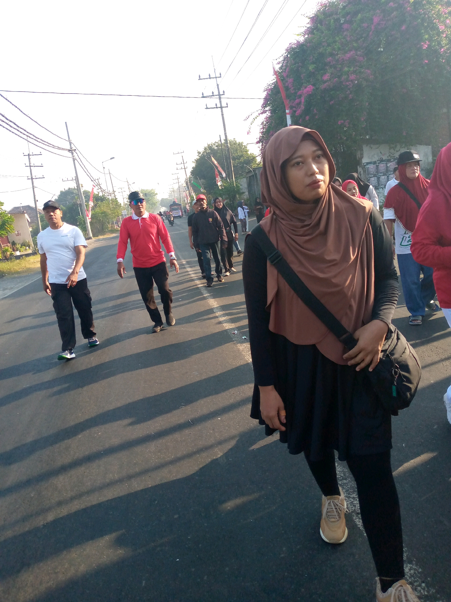 In Sumberwono, Indonesia, a group of people are walking down a paved road under a bright, hazy sky. A woman wearing a brown hijab and a black dress, with a black bag slung across her shoulder, walks towards the viewer in the foreground. Behind her, several men are walking away from the viewer. Two men in the middle ground are wearing white t-shirts and black pants, and red long-sleeved shirts and black pants, respectively. Further in the background, more people are visible, some wearing red shirts and hats. The road is lined with trees and some buildings, and there are utility poles with wires stretching overhead. Red and white Indonesian flags are visible on poles along the side of the road. The sun casts long shadows across the pavement, indicating it is likely morning or late afternoon.