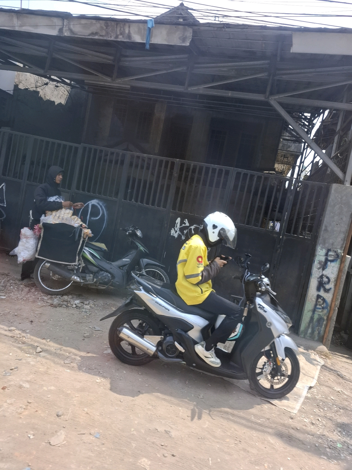 Two individuals on motorcycles are present in a street scene. The first individual, wearing a dark hooded jacket and black pants, stands next to a black motorcycle with a large black storage box on the rear, filled with what appears to be packaged goods. A white plastic bag is also attached to the motorcycle. This motorcycle is parked on a dirt and gravel surface, with a black metal gate and a building structure visible in the background.
The second individual, seated on a silver and black motorcycle, wears a bright yellow jacket, black pants, and a white helmet with a clear visor. This individual is holding a black object, possibly a smartphone, in their hands. The motorcycle is positioned on a lighter colored surface, with a concrete pillar and graffiti visible to the right. Both motorcycles appear to be parked or stopped. The environment suggests a street or alleyway in an urban setting. The overall lighting indicates it is daytime, with shadows cast on the ground. The location is identified as Sukasari, Indonesia.