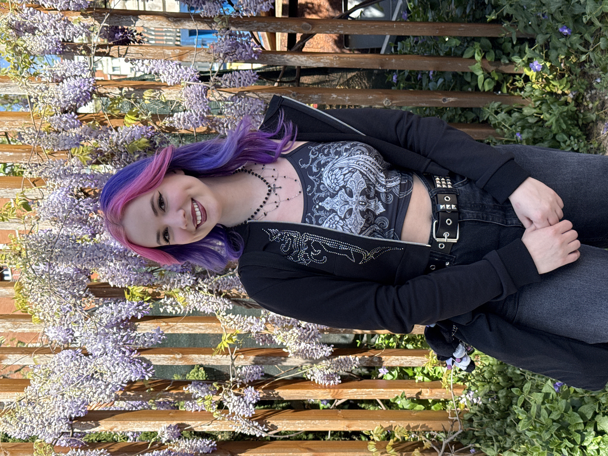 A young woman with dyed purple and pink hair stands smiling in front of a wooden trellis covered in wisteria flowers and some green foliage. She wears a black zip-up jacket with silver embellishments, a grey patterned top, black jeans held with a studded belt, and a beaded necklace. The trellis is made of horizontally stacked wooden planks. Buildings can be seen faintly through the trellis in the background. The location is unknown and is NOT identified as Madrid, Spain.