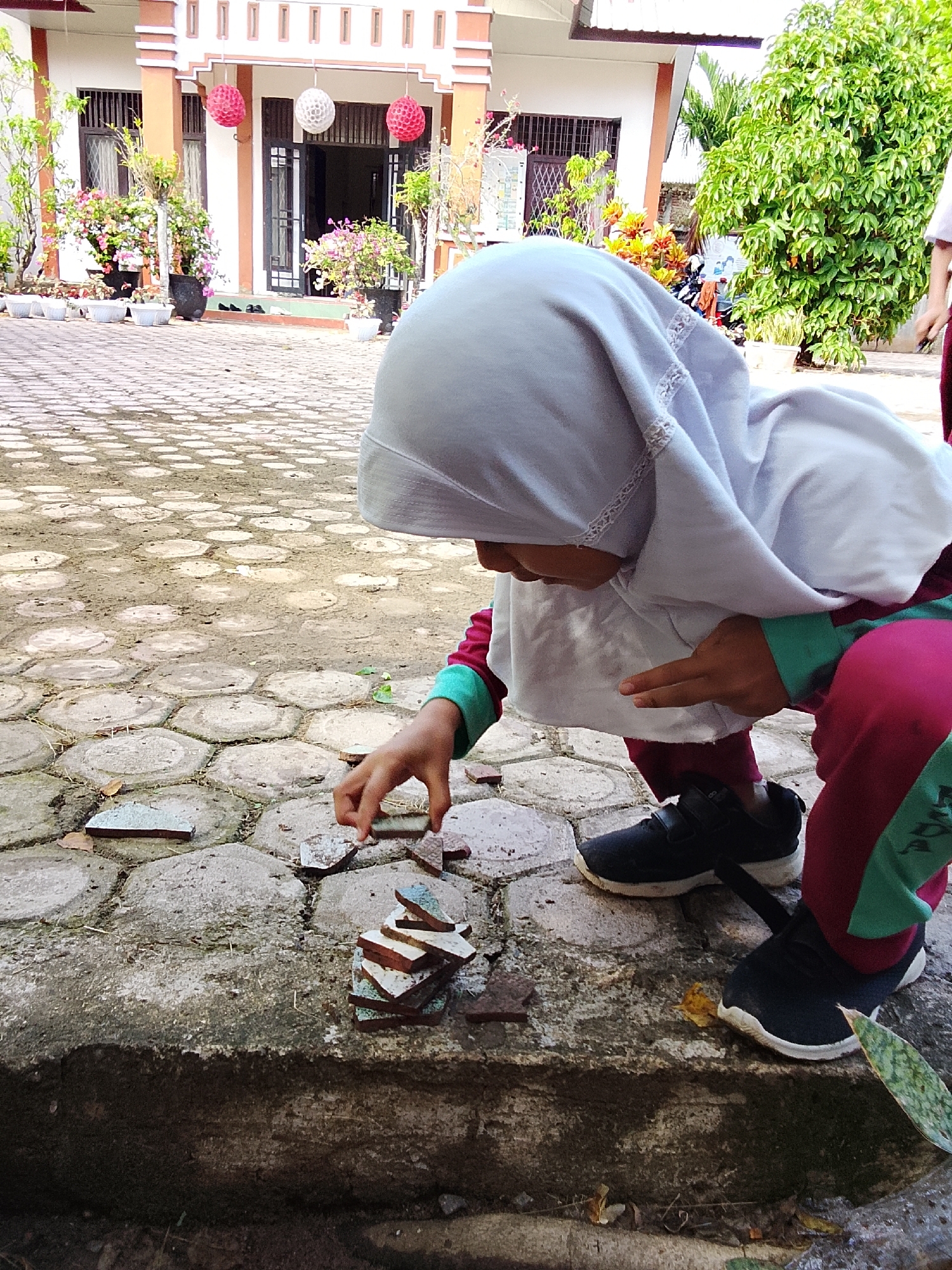 The image depicts a young child wearing a white hijab and colorful clothing, crouched down outdoors and playing with broken ceramic pieces. The child is stacking the ceramic fragments on a concrete ledge. In the background, a building with an orange and white facade and several potted plants are visible. The ground is paved with hexagonal stones. The scene is likely located in Keudejungkagajah, Indonesia, given the architectural style and vegetation. The child's attention is focused on the task at hand, demonstrating a solitary activity. The lighting suggests it is daytime.
