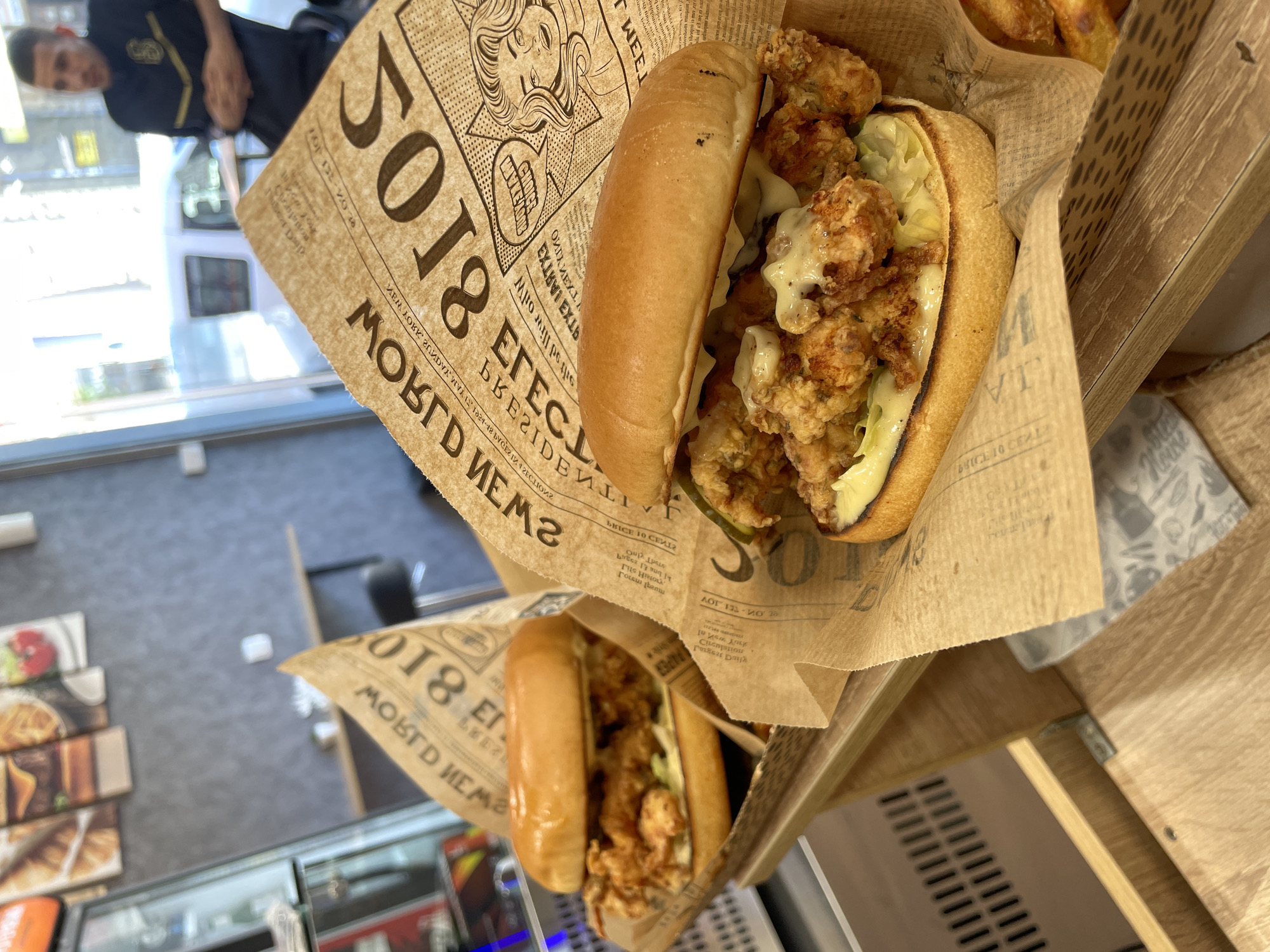 Two fried chicken burgers are presented on wooden trays, each wrapped in newspaper-style paper. The burgers have golden-brown buns and are filled with crispy fried chicken, lettuce, and sauce. In the background, a man wearing a dark shirt with gold accents stands near a window, and there are glimpses of restaurant interior elements like a glass display case and shelves. The scene is likely a fast-food establishment or cafe. The image is taken from a slightly elevated angle.