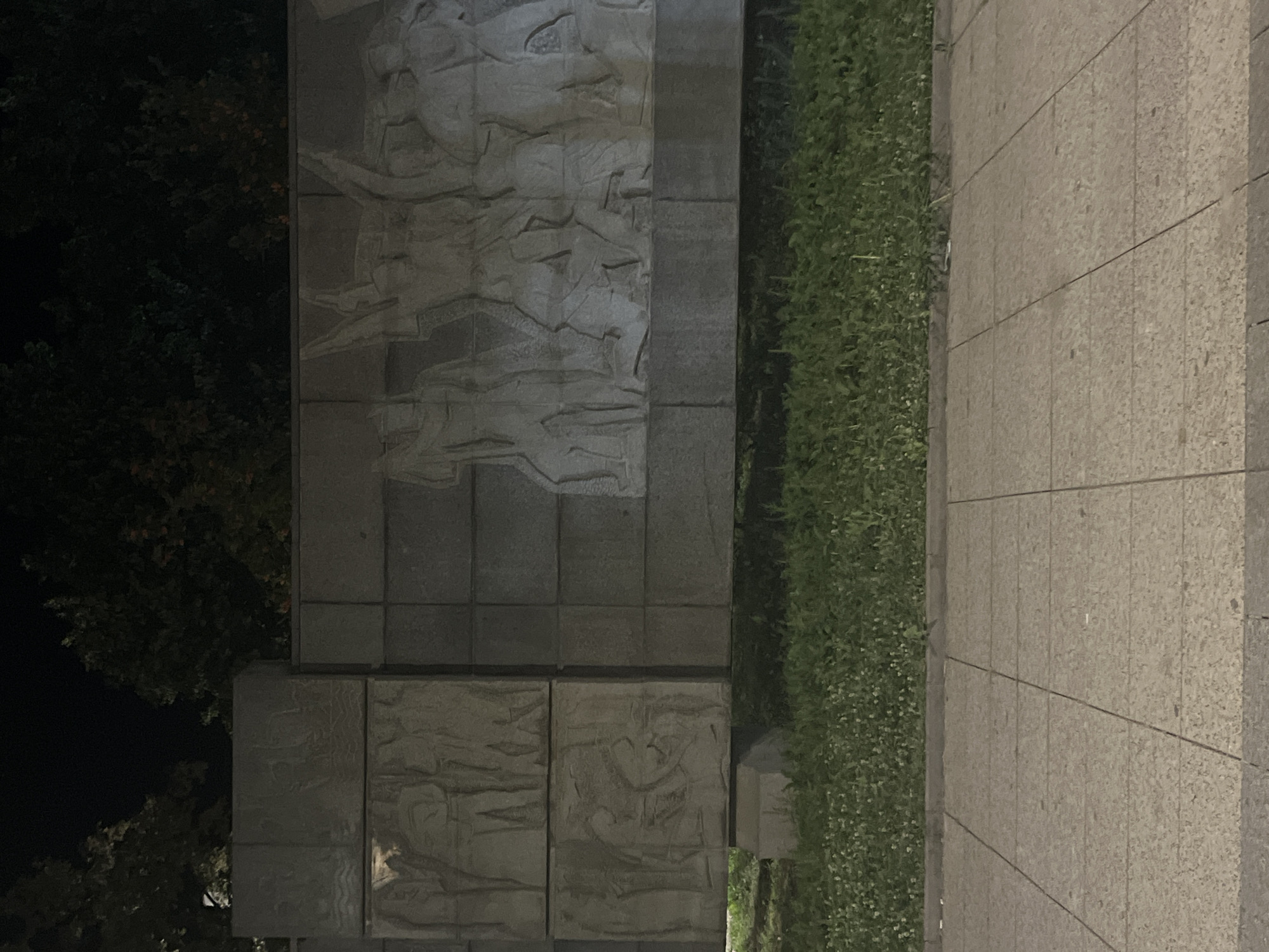 The image captures a nighttime view of a bas-relief monument located in Vratsa, Bulgaria. The monument is constructed from large, rectangular stone blocks, with carved figures depicting a historical or allegorical scene. The relief features multiple human figures in various poses, some appearing to be in motion or engaged in an activity. The foreground of the image shows a paved walkway made of light-colored square tiles, with a strip of green grass bordering the monument. Trees with dark foliage are visible in the background, silhouetted against the night sky. The lighting suggests artificial illumination, casting shadows and highlighting the textures of the stone carvings.