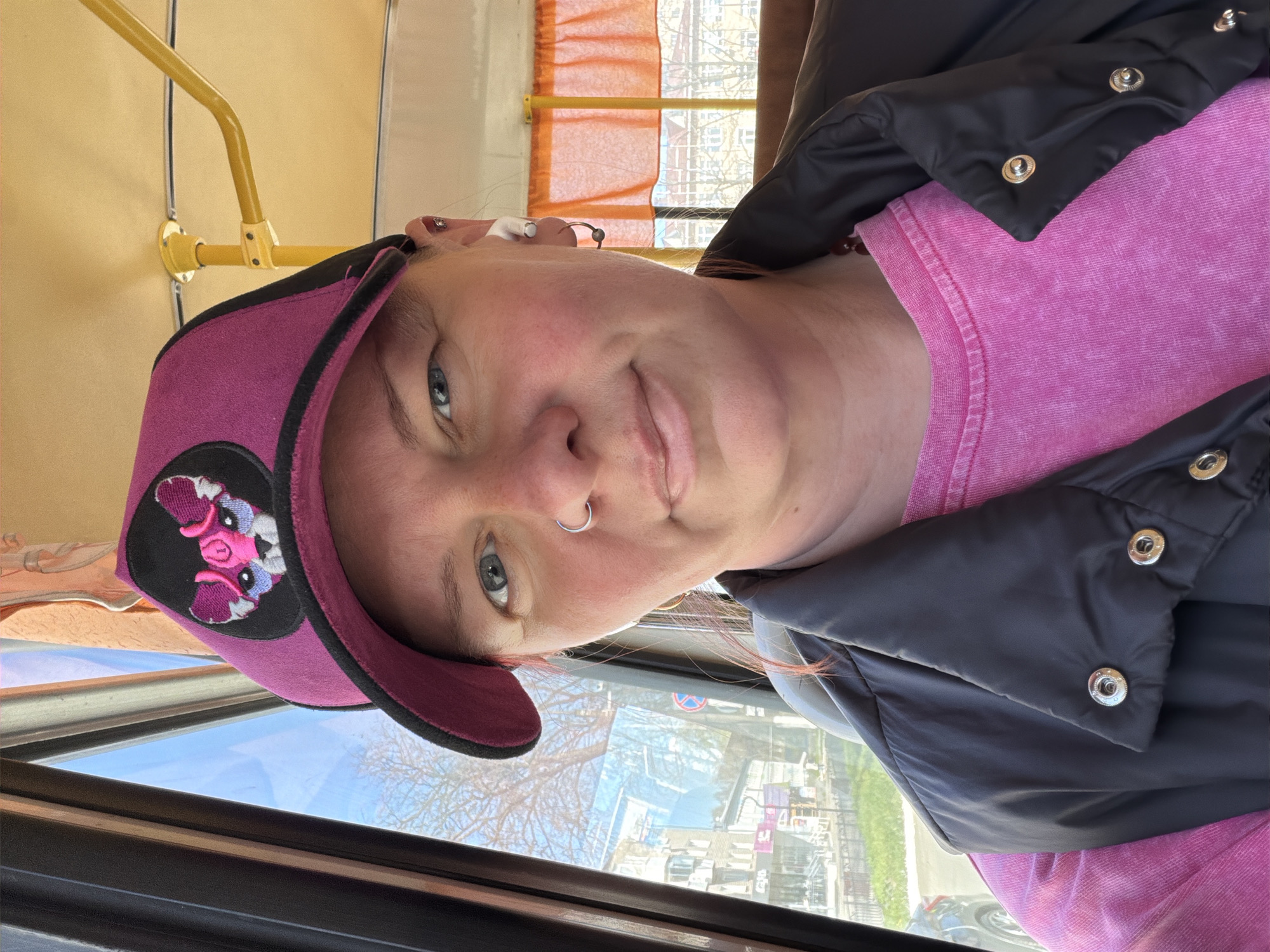 A fair-skinned woman with blue eyes and a nose ring, wearing a pink t-shirt, a black jacket, and a pink baseball cap with a cartoon pig patch, is seen from a close-up, slightly skewed perspective inside a bus or tram. The environment outside the window shows buildings, trees without leaves, and what appears to be the reflection of a city, likely Chaikovsky, Russia, based on the presence of buildings, infrastructure, and foliage that could be found in that region. Parts of the interior of the bus/tram, including a yellow support pole, are also visible.