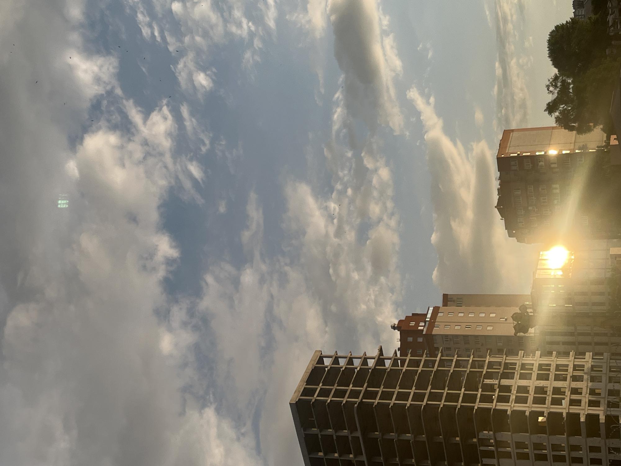 The image shows a city skyline at sunset, with the sun casting a warm glow on the buildings. In the foreground, an unfinished concrete building stands tall, its skeletal structure exposed. To its right, a completed residential building with numerous windows is visible. Further in the distance, another residential building with an orange facade is seen, with the sun reflecting off its windows. The sky is filled with scattered clouds, and several birds are seen flying. The scene appears to be located in the city of Dnipro, Ukraine, as indicated by the architectural style and the overall urban landscape. The lighting suggests a warm, late afternoon or early evening time.