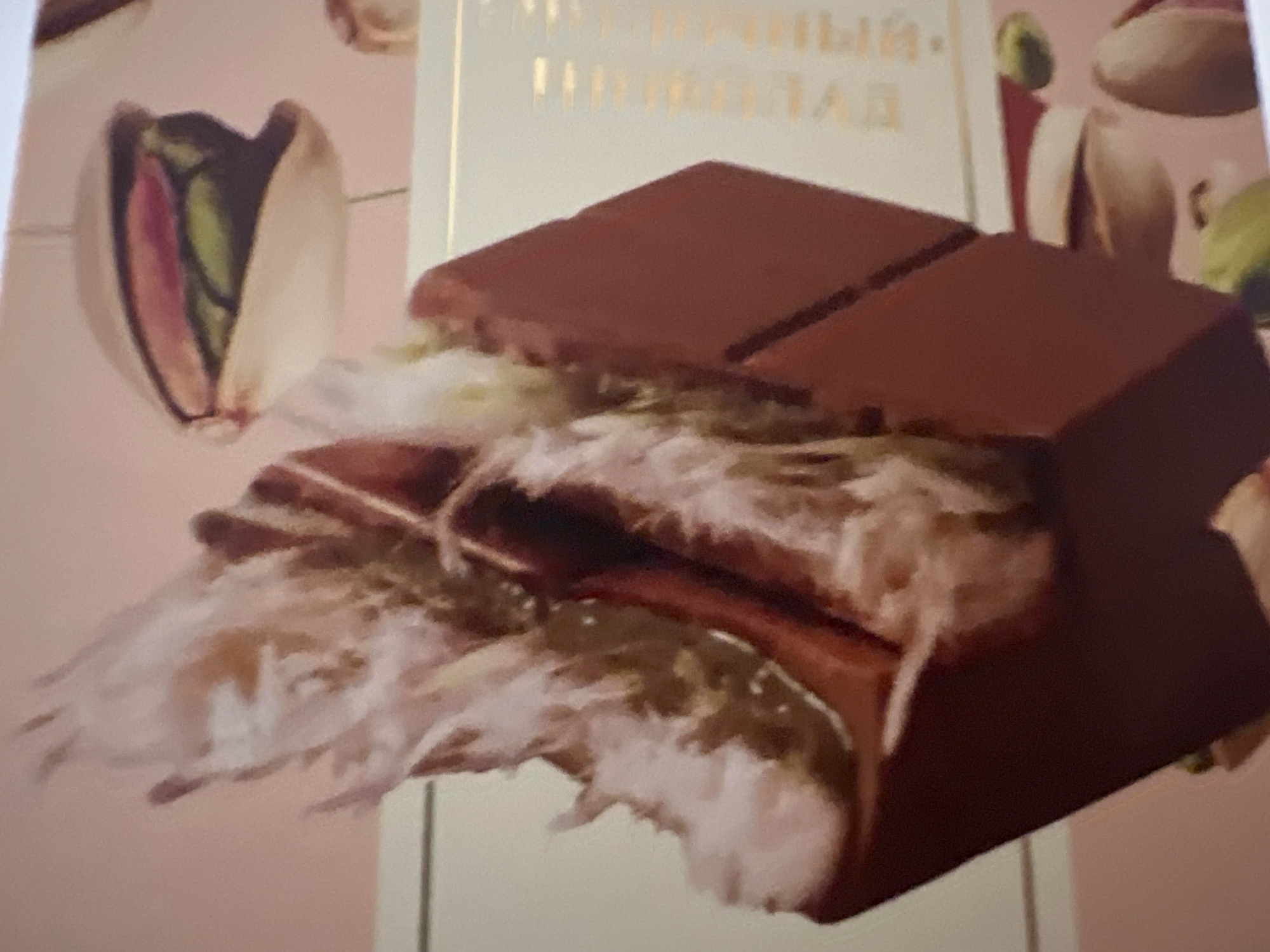 The image depicts a close-up, slightly out-of-focus view of a confectionery item, likely a chocolate bar with a pistachio and fluffy filling. The chocolate appears to be dark brown and has a geometric, almost block-like structure. Emerging from the chocolate is a wispy, light-colored filling, possibly nougat or a similar confection, with visible strands. In the background, there are blurred images of pistachios in their shells, with some showing the distinct green and purple interiors. The overall color scheme is dominated by pink and light beige, suggesting a product package or a stylized advertisement. The lighting is soft and diffused. There are no people or signs of a specific location within Yekaterinburg, Russia visible in this image; it appears to be a commercial product shot.