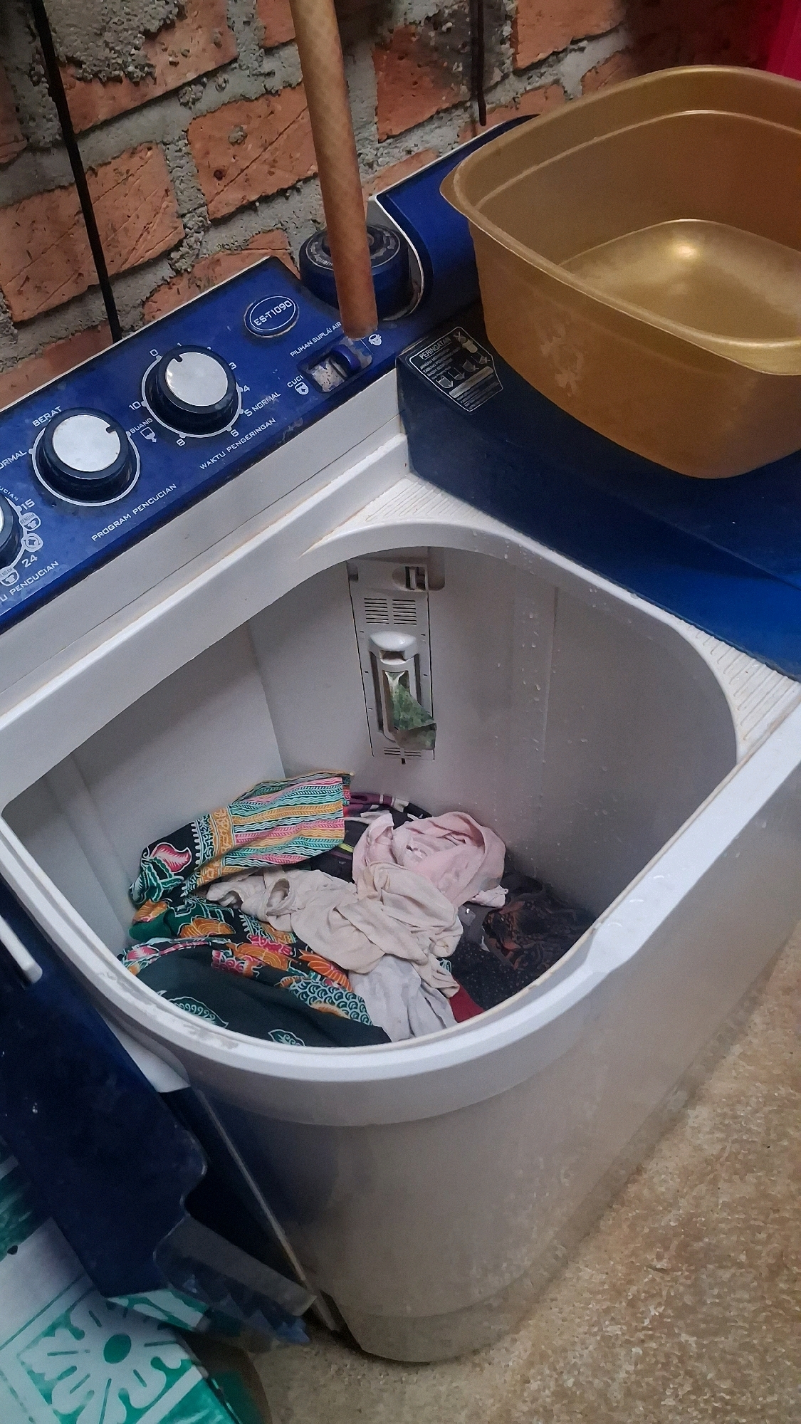 The image shows a top-load washing machine with a basket of laundry inside. The washing machine is blue and white and has several control knobs and buttons on the top. A golden plastic basin sits on top of the washing machine, to the right of the controls. The background appears to be a brick wall. The scene is likely located in a residential setting in Karang Sari, Indonesia, as indicated by the presence of the washing machine and the typical brickwork seen in Indonesian homes. The washing machine appears to be in use or recently used, as indicated by the laundry inside. There are no people visible in the image.