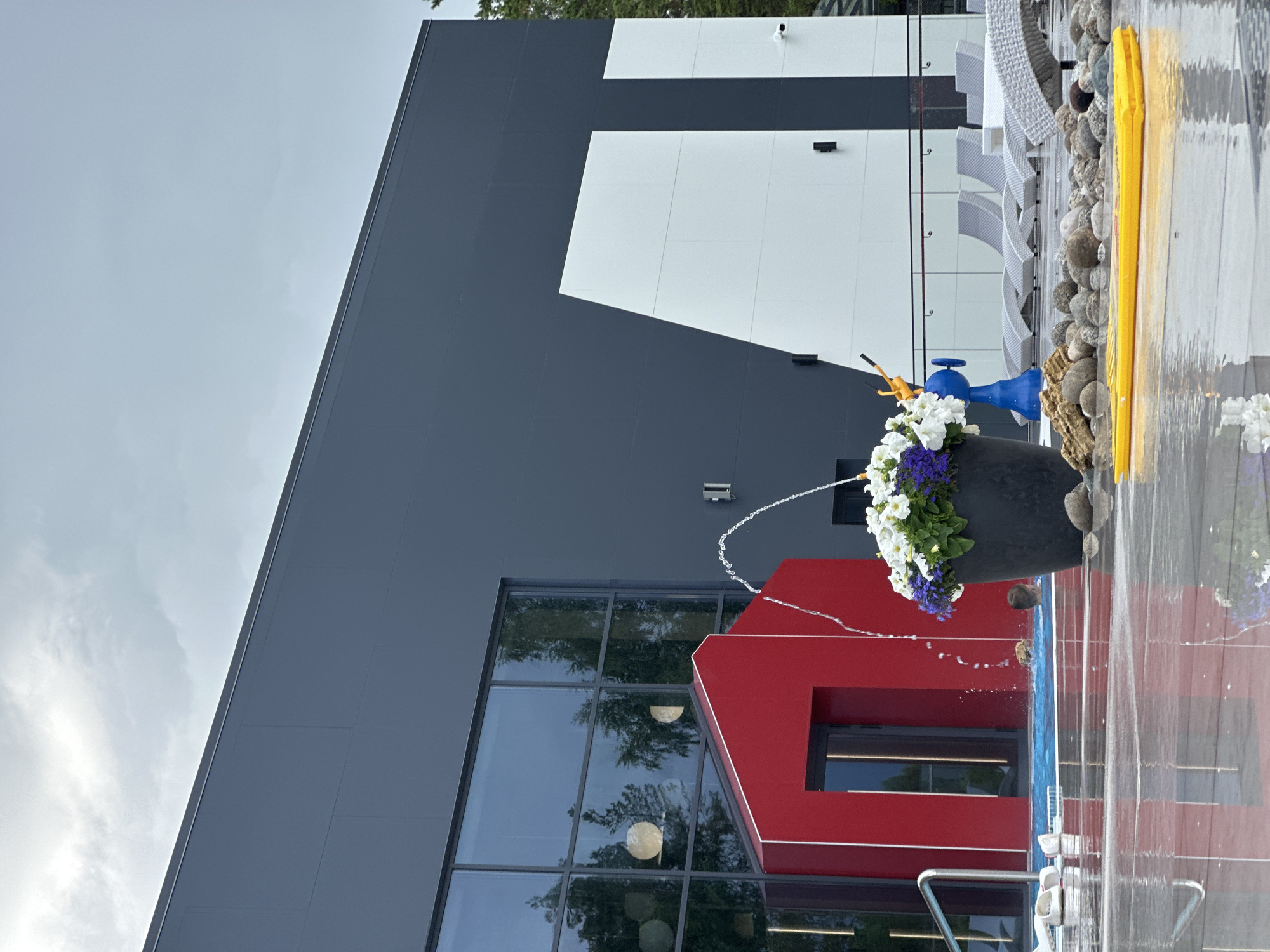 The image shows a modern building with a dark gray facade and large white geometric shapes. A bright red architectural element juts out from the building's lower section. In the foreground, there is a black planter filled with white and purple flowers, with a blue fountain spout spraying water upwards. To the right, a yellow object, possibly a life preserver, rests on a surface covered with pebbles. A pool or water feature is visible with water cascading down, creating a transparent curtain. The overall scene appears to be outdoors, with hints of trees reflected in the building's windows. The sky is overcast, suggesting a cloudy day. The orientation of the image is rotated 90 degrees counter-clockwise. No people are visible in the image, and no text is discernible.