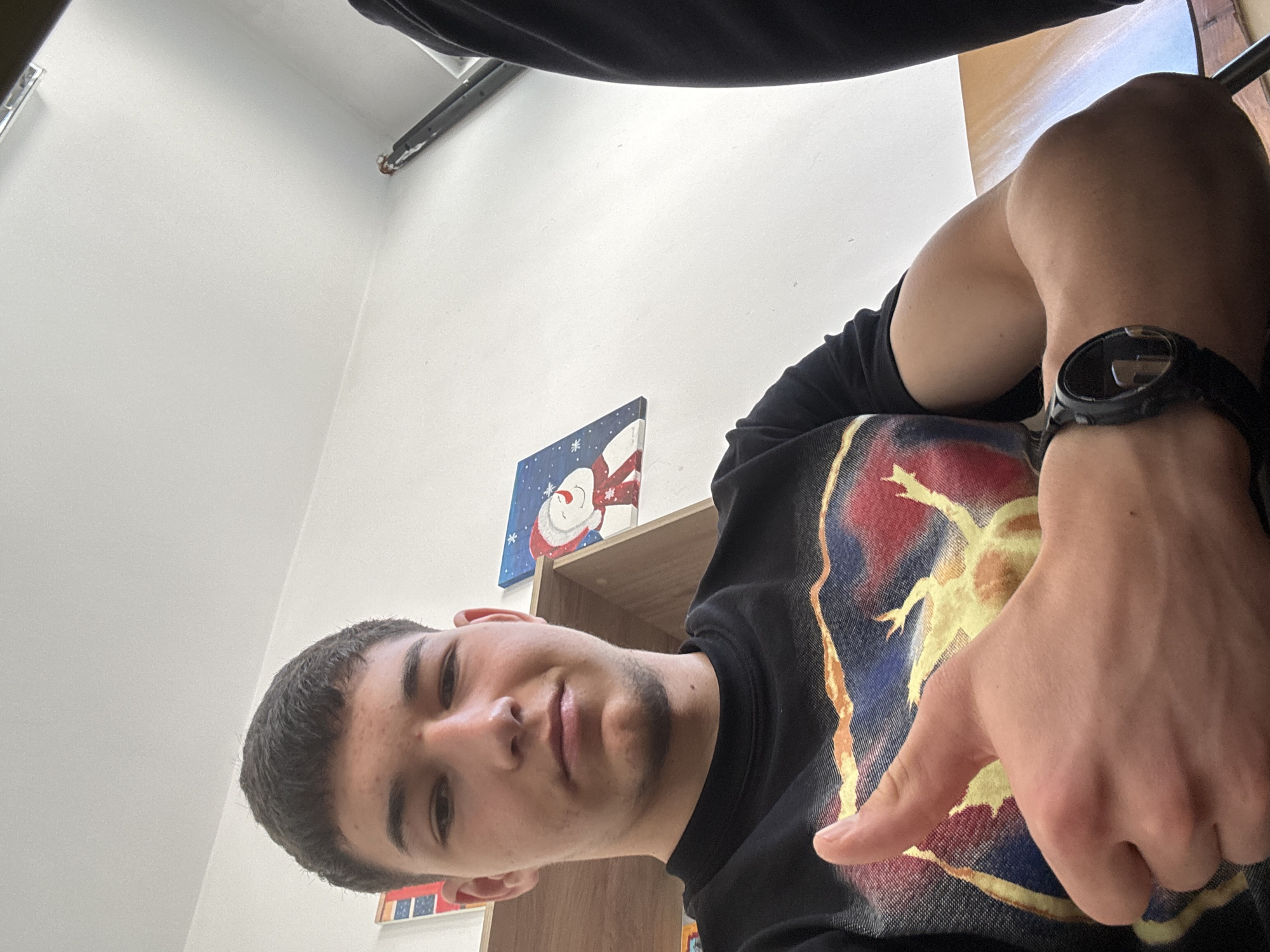 A young man is taking a selfie indoors. He is wearing a black t-shirt with a colorful design and a black watch on his left wrist. He is pointing at the t-shirt with his right hand. Behind him is a white wall and a wooden shelf. A painting of a snowman is on the shelf. It is not possible to determine the location in Zlatograd, Bulgaria, from the provided image.