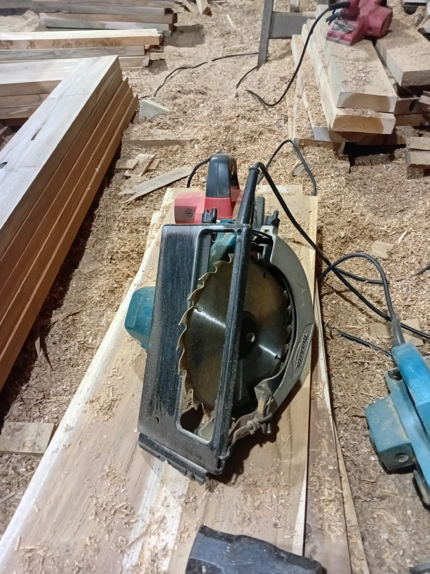 In a workshop filled with sawdust, a circular saw rests on a wooden plank. The saw, partially covered by wood shavings, has a silver blade with black teeth. To its left, a stack of lumber is neatly arranged. In the background, another power tool, possibly a sander, is visible, along with more stacks of wood and scattered sawdust. The scene suggests ongoing woodworking activities. This location is the craftsman's workshop in Playen, Gunungkidul Regency, Yogyakarta Special Region, Indonesia.