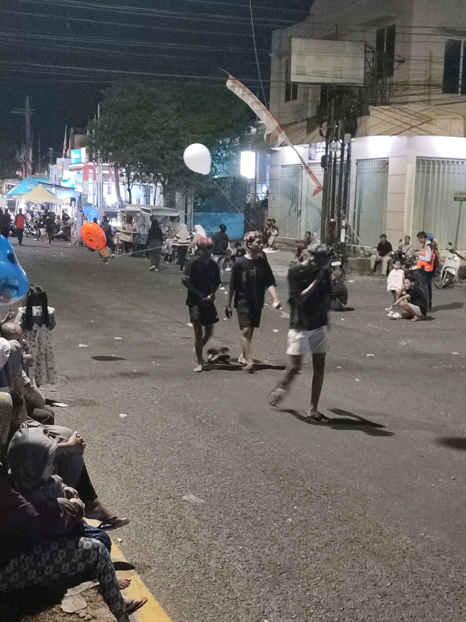 The image depicts a nighttime street scene in Jember, Indonesia, likely during a festive event or market. Several people are walking and standing on a paved road. In the foreground, to the left, several individuals are seated along the edge of the road, some appearing to be children.
In the mid-ground, three young men are walking towards the camera. The one on the left is wearing a dark t-shirt and shorts, with light-colored hair. The middle individual is also in dark clothing, and the person on the right is wearing a dark t-shirt and white shorts, carrying what appears to be a camera or filming device.
Further back, there are various stalls and tents set up, suggesting a temporary marketplace or event area. A white balloon is visible in the upper left portion of the image, and an orange balloon is floating in the mid-ground. There are also buildings and streetlights visible in the background, indicating an urban environment. The overall lighting suggests it is dark, with artificial lights illuminating the scene. The ground is littered with some debris.