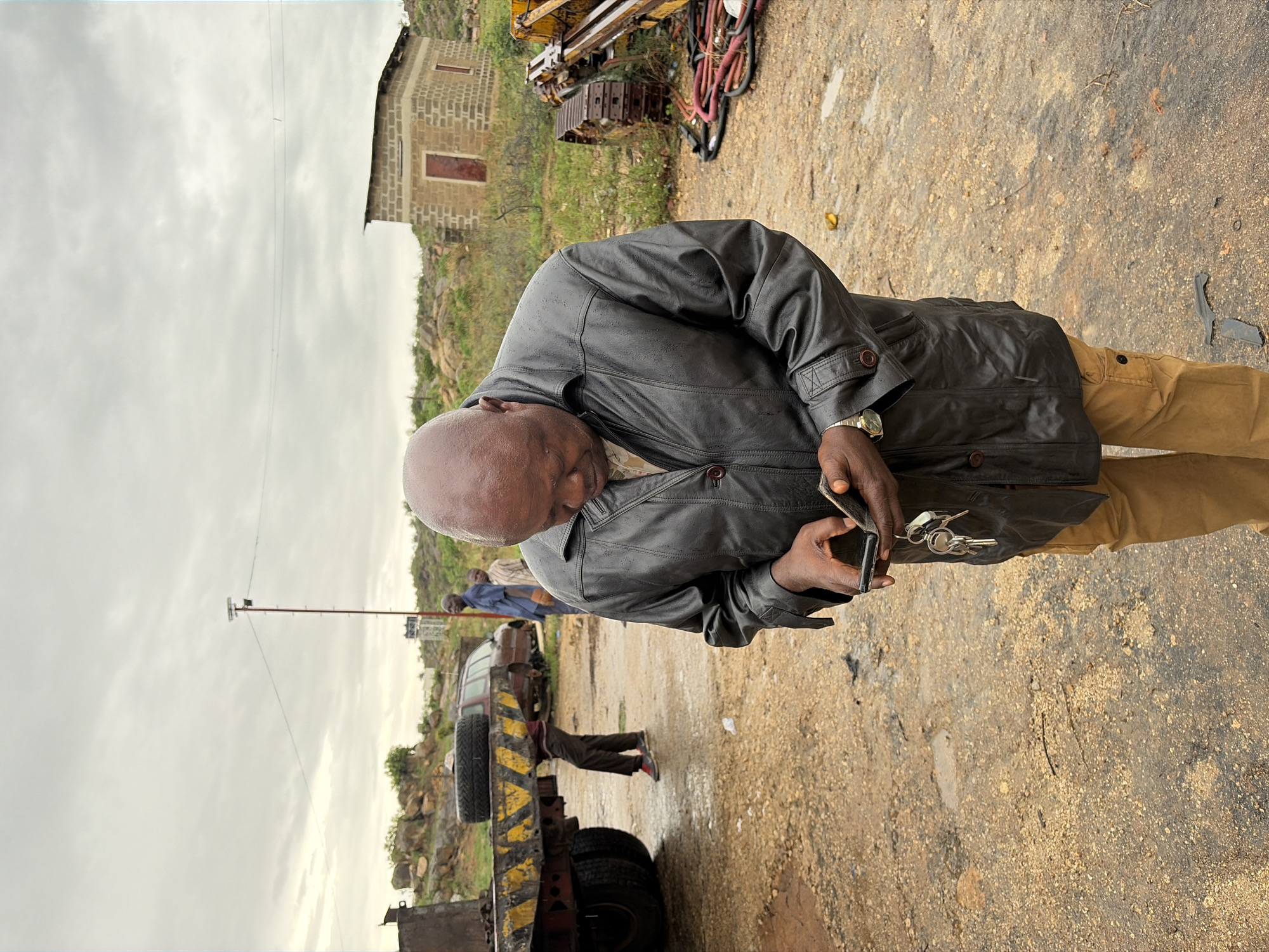 A medium shot of an older bald man in a black leather jacket and khaki pants looking down at his phone and holding a wallet with keys in his left hand. He is wearing a gold watch. The ground appears to be dirt and gravel. In the background is a truck with a tire mounted on the side, and a person working underneath the truck. Further back are two other people and a light pole. To the far left, a stone structure can be seen. In the background, there are rocks and some sparse vegetation. In the far upper left, there is some construction equipment and coiled hoses. The sky is overcast. Based on the prompt, the location is Gura Top, Nigeria.