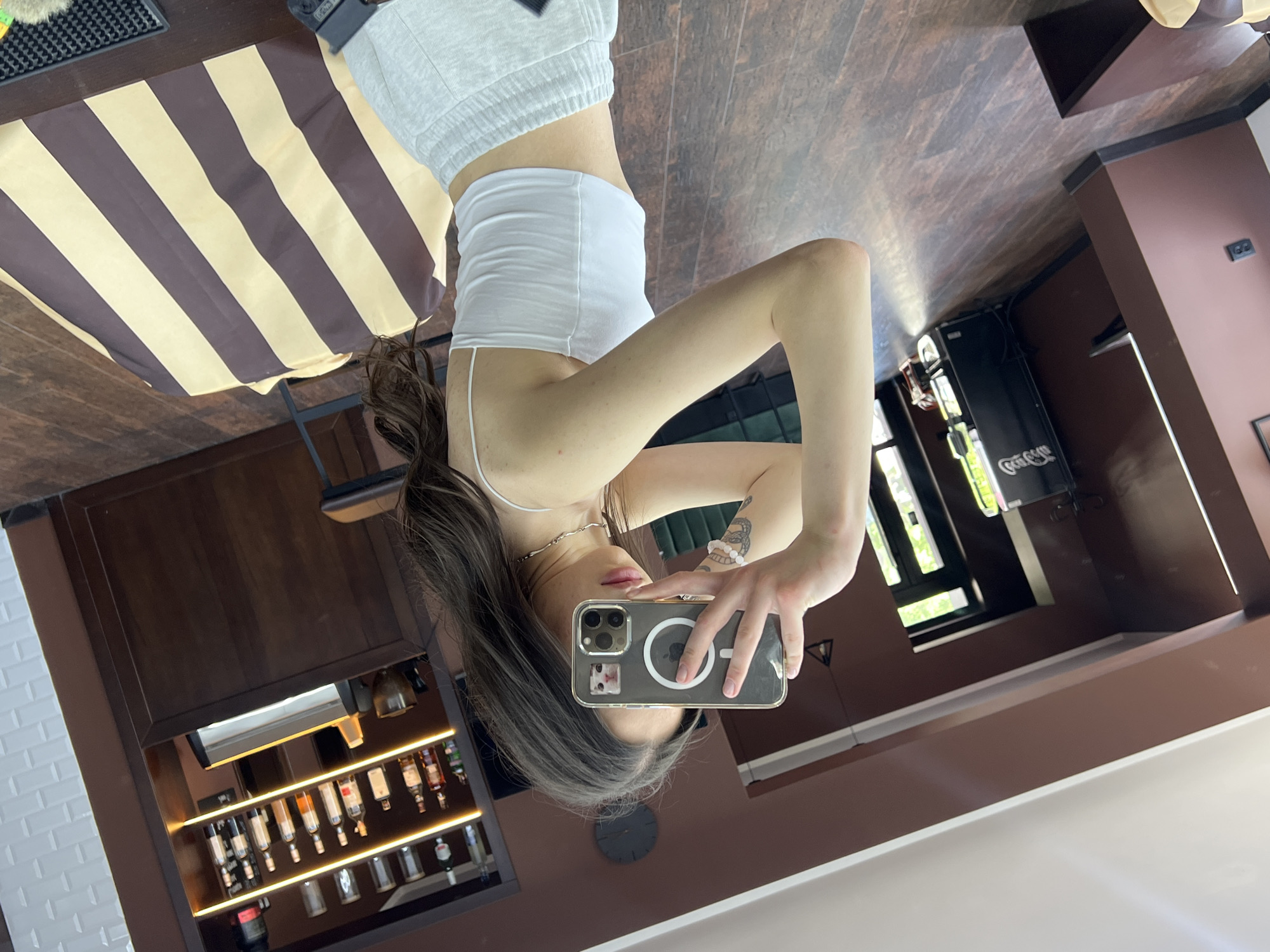 A young woman takes a selfie in what appears to be a cafe or bar in Ternopil, Ukraine. She is wearing a white tank top and gray shorts. A striped awning is partially visible in the background, along with shelves of bottles and glasses. The lighting is bright. A Coca-Cola refrigerator is also visible.