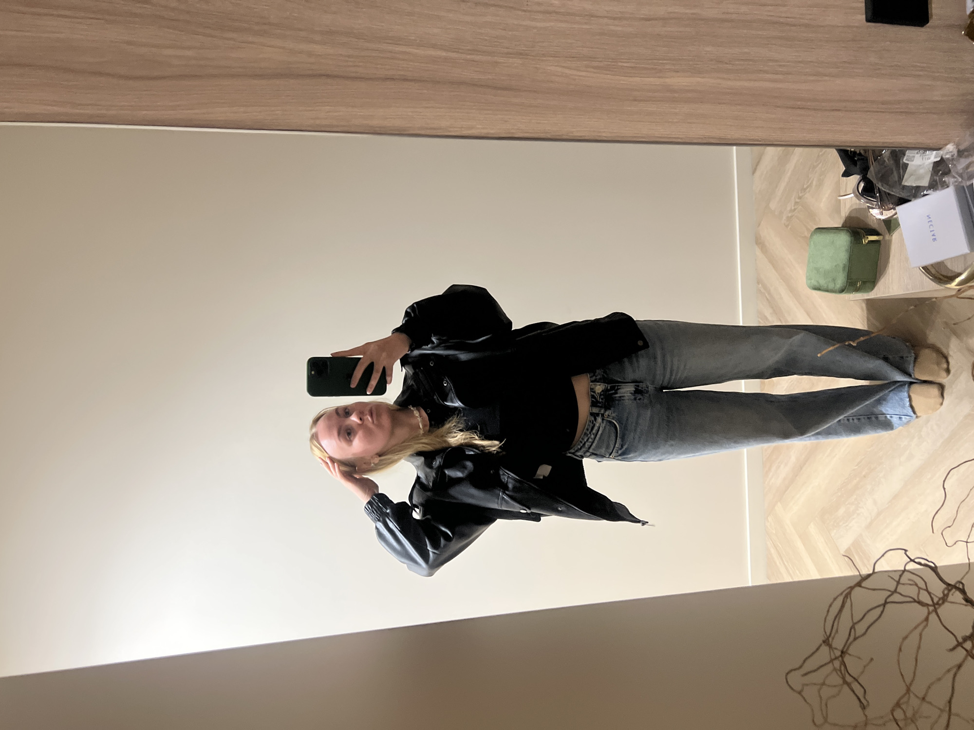 A young woman stands indoors, taking a mirror selfie with a smartphone. She is wearing a black leather jacket, jeans, and has blonde hair. A small green jewelry box and a white box are visible on a surface to the right of her. The location is unknown and cannot be determined from the image.