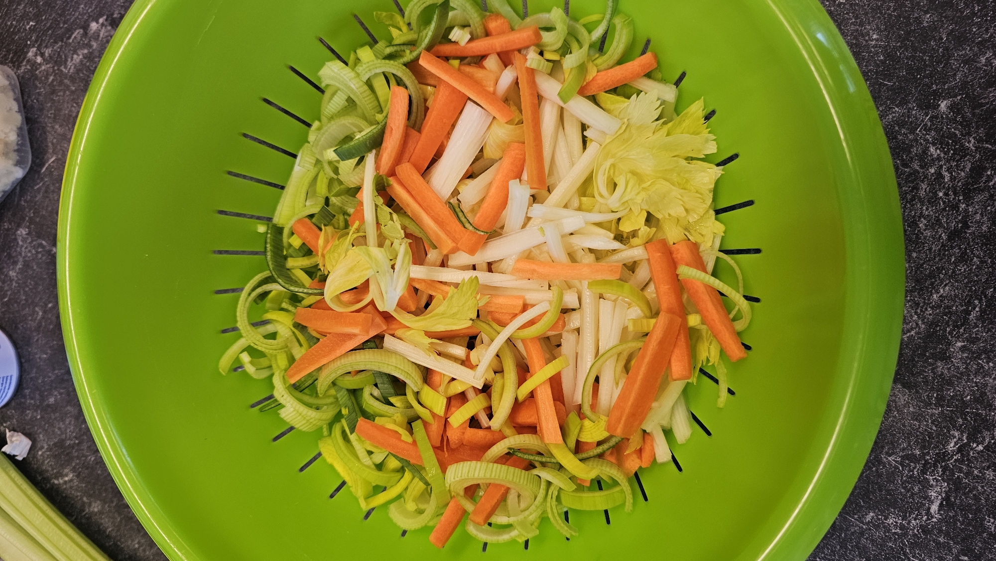 An overhead shot captures a vibrant green colander filled with an assortment of freshly chopped vegetables, resting on a dark gray, speckled countertop. The colander's inner surface features small, elongated slots. The main subjects are the mixed vegetables, meticulously cut into uniform pieces.
Dominating the mix are bright orange carrot sticks, appearing julienned or in small batonnets. Interspersed among these are light green and white rings or half-rings of what appears to be sliced leeks, and pale white, thin stalks of celery, some still retaining small, yellow-green celery leaves. The various colors of the vegetables create a visually appealing contrast within the green colander.
The activity depicted is the preparation of ingredients, as the vegetables are clean and uniformly chopped, suggesting they are ready for cooking or washing. To the top-left of the colander, a blurry, light-colored object, possibly a container, is partially visible. In the bottom-left corner, a single, unchopped stalk of celery can be seen, partially off-frame. No visible text is present in the image. The lighting is bright and even, illuminating the scene clearly.