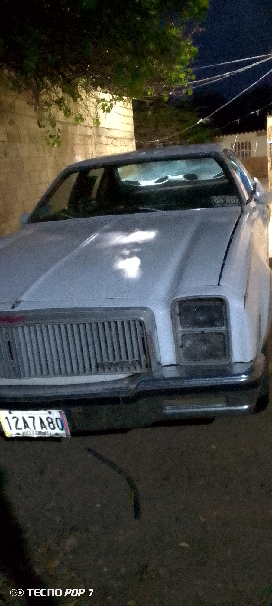 The image shows the front of a white car parked on a paved surface at night. The car has a prominent vertical grille and rectangular headlights. A license plate with the characters "12A7A80" is visible on the front bumper. The background includes a light-colored brick wall, some foliage from a tree, and utility wires against a dark sky. The scene appears to be in an urban or semi-urban environment in Altagracia, Venezuela, based on the presence of a license plate from that region, likely on a street or driveway. The lighting suggests artificial light sources are present, possibly from streetlights or nearby buildings. There are no people or clear signs of recent activity in the frame.