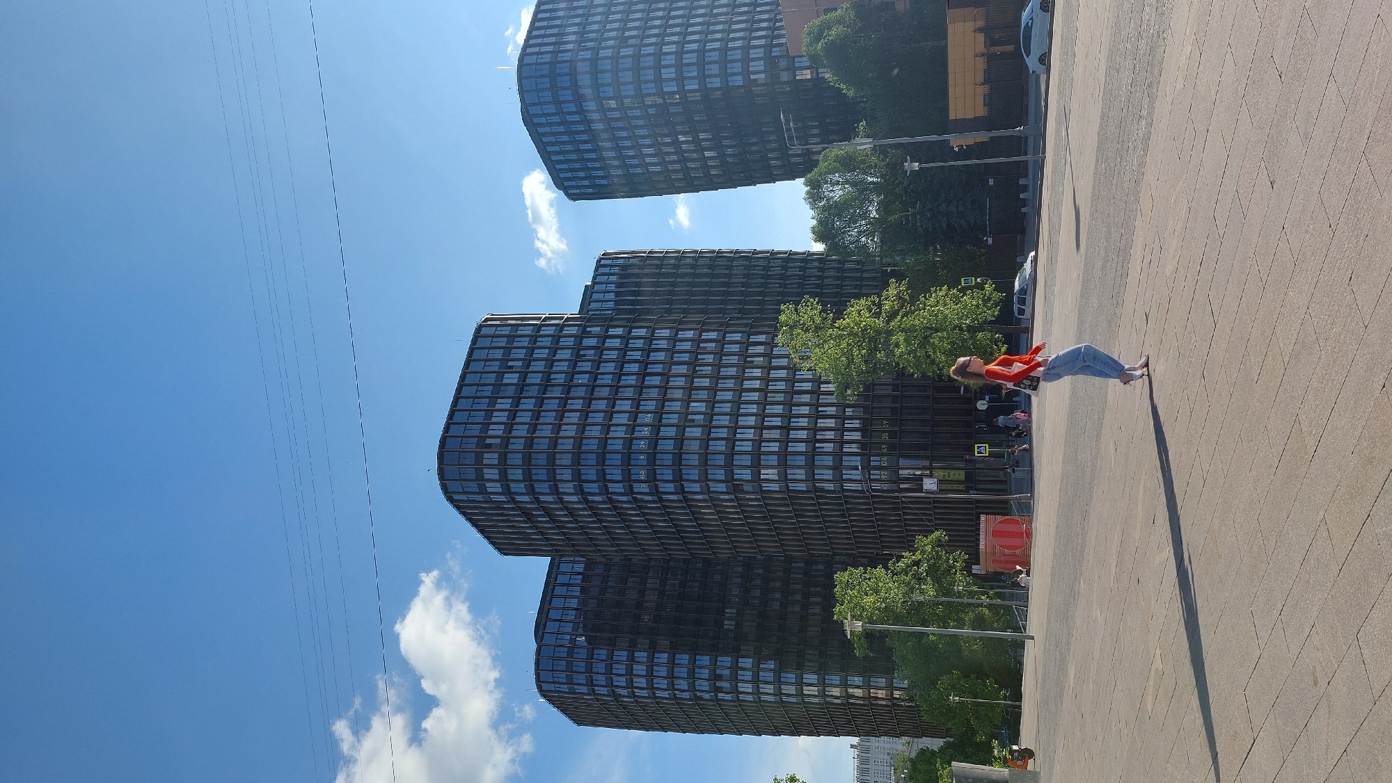 The image shows a paved square with three large, dark-colored office buildings with a mirrored-window design in the background. The sky is blue with some clouds. In the foreground, a woman wearing a red shirt and jeans is walking towards the buildings. The location appears to be in Moscow, Russia, judging by the architecture of the buildings. There are trees and some other people visible near the buildings.