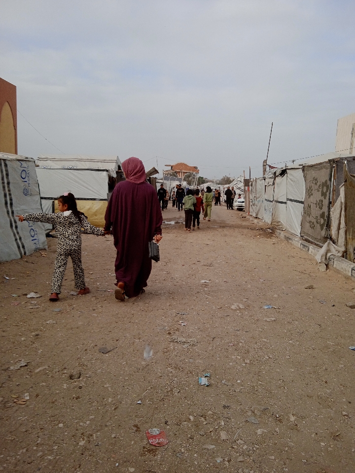 A woman in a maroon headscarf and robe walks with a young girl in a leopard print outfit down a dirt road. The girl points to the left. They walk away from the camera, heading toward a group of people further down the road. Makeshift shelters line both sides of the road. Litter and debris are scattered on the ground. The sky is overcast.