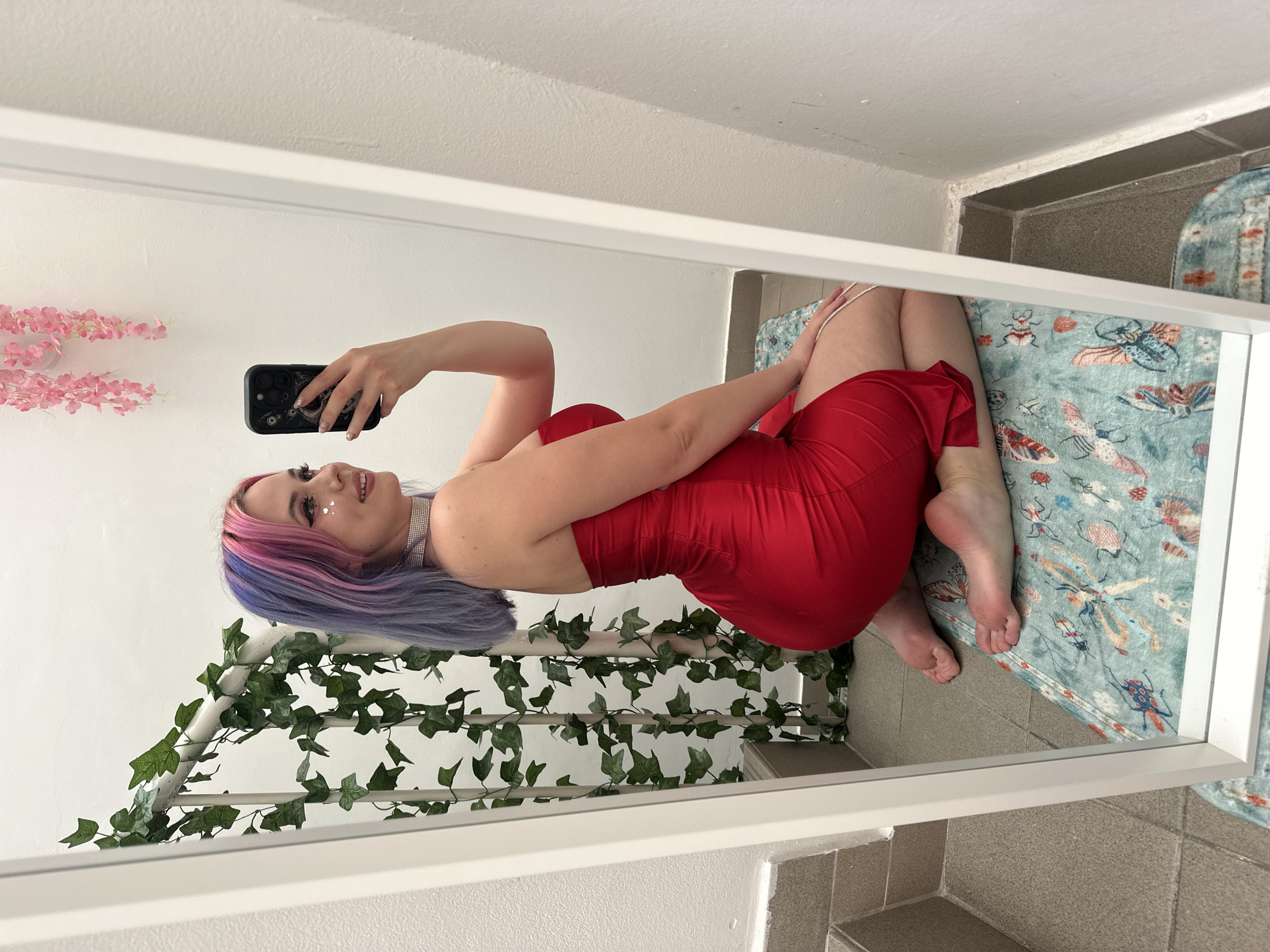 A reflection in a white-framed mirror shows a woman in a red dress posing and taking a selfie. She is kneeling on a rug with insect designs. The wall behind her is decorated with ivy and pink flowers. She has pink and purple hair and is wearing a choker. The room features beige tile flooring and what appears to be a white wall and ceiling. Based on the limited information, it is not possible to confirm that this is in Zagreb, Croatia.
