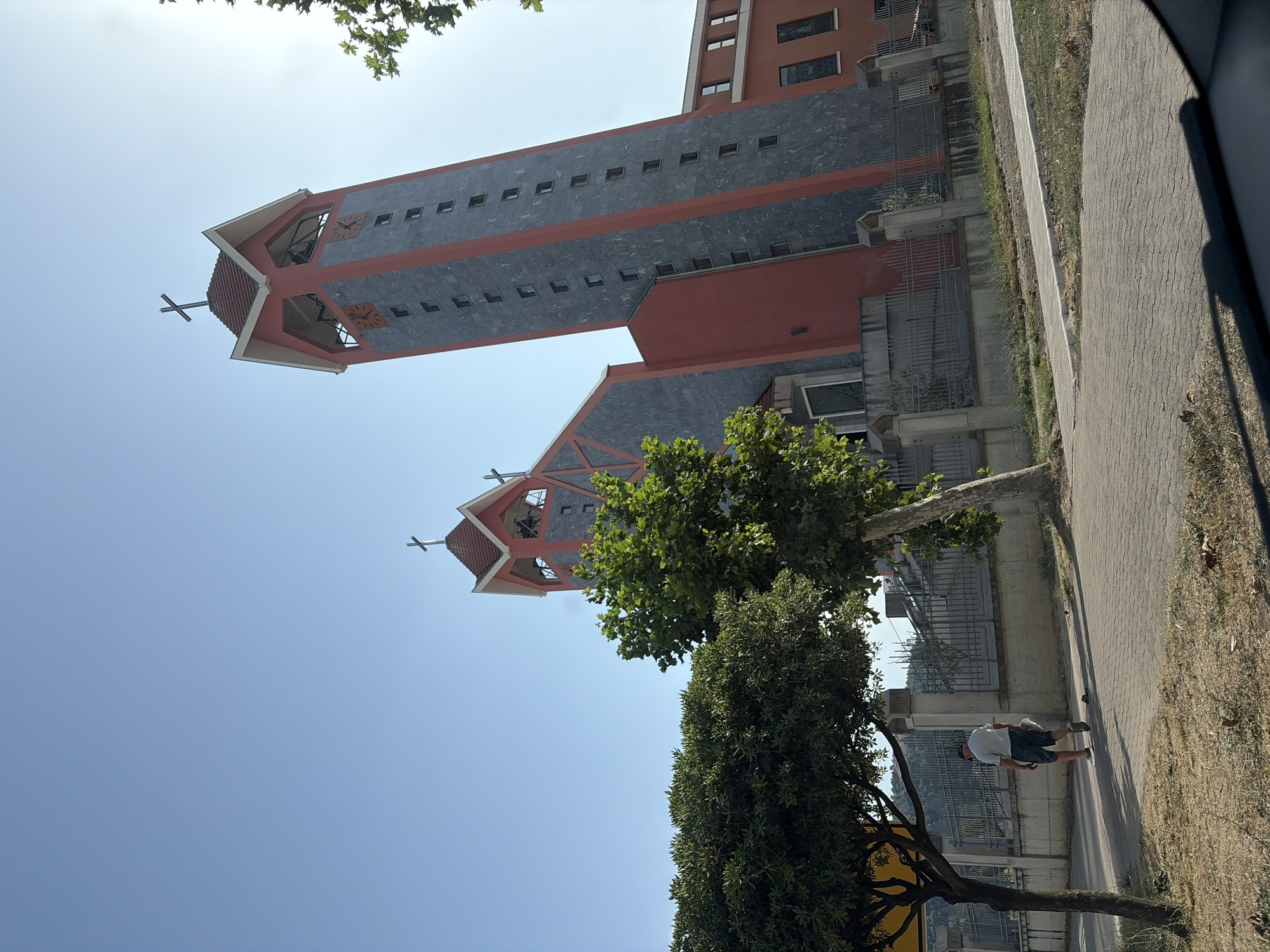 The image shows a modern church building with two tall towers, one significantly taller than the other, against a clear blue sky. The church has red and grey stone exterior. A cross is visible atop each tower. In the foreground, a man in a light-colored shirt and dark shorts walks away from the camera on a paved walkway. He is walking past a sidewalk and a fenced area. Trees with green foliage are present in the foreground and to the right of the church. The environment appears to be urban, with a fence and part of a building visible in the background. The scene is captured on a sunny day.