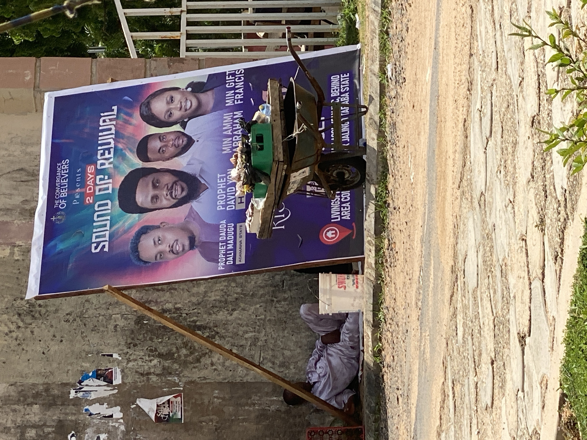 This media file depicts an outdoor scene in Jalingo, Nigeria, likely during the daytime given the bright sunlight. The focal point is a large banner advertising a "SOUND OF REVIVAL" event presented by "THE CONVERGANCE OF BELIEVERS," scheduled for "2 DAYS." The banner features portraits of several individuals, including "PROPHET DAUDA DALI MADUGU," "PROPHET DAVID YON," "MIN AMMI ABRAHAM," and "MIN GIFT FRANCIS." Location details such as "ADAMAWA STATE" and "JALINGO 1 TAF ABA STATE" are also visible on the banner.
Adjacent to the banner is a rustic wheelbarrow filled with various items, including what appears to be trash and a green crate. A man, dressed in light-colored traditional clothing, is seen resting or sleeping in the shade provided by the banner, leaning against a wall. A white bucket with the brand name "SWAN" is placed near him. The setting appears to be a somewhat neglected urban or semi-urban environment, with a rough, uneven ground surface and scattered debris. In the background, a metal fence and some greenery can be observed. The overall impression is one of everyday life, with a religious event poster juxtaposed with elements of informal commerce or street life.