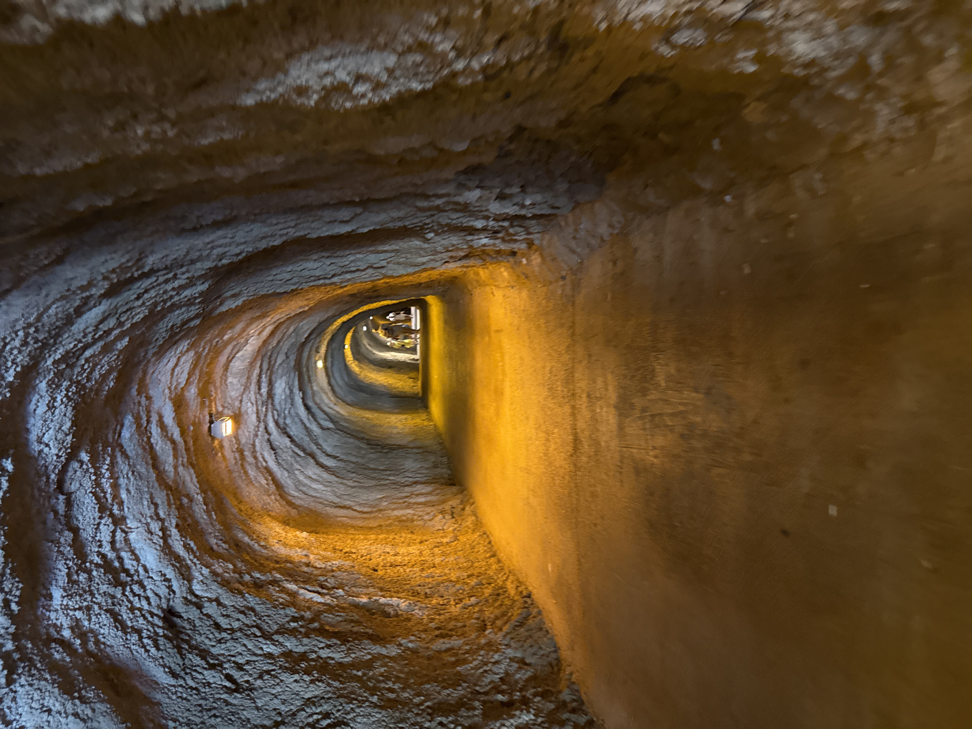 An image taken from inside a tunnel in Kamenovo, Montenegro shows a rough-hewn, curved passageway. The walls and ceiling are textured, appearing to be made of rock or compacted earth. The tunnel is illuminated by artificial lighting, casting a warm, orange glow on the pathway and the rough surfaces of the tunnel. A few figures are visible in the distance, walking towards the light at the far end of the tunnel. The ground is a smooth, packed dirt or gravel surface. The overall impression is one of an ancient or natural pathway enhanced with lighting for passage.
