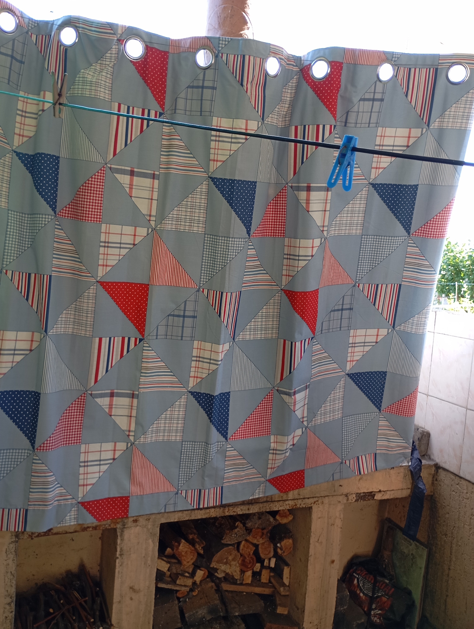 In Horishni Plavni, Ukraine, a patterned shower curtain with a geometric design of triangles in blue, red, and white, hangs from a clothesline secured by two clothespins. The curtain is suspended in an outdoor setting, likely a backyard or balcony, with glimpses of greenery and a tiled wall in the background. Below the curtain, a recessed area contains stacked firewood, suggesting an outdoor utility space. The overall scene is illuminated by natural daylight.