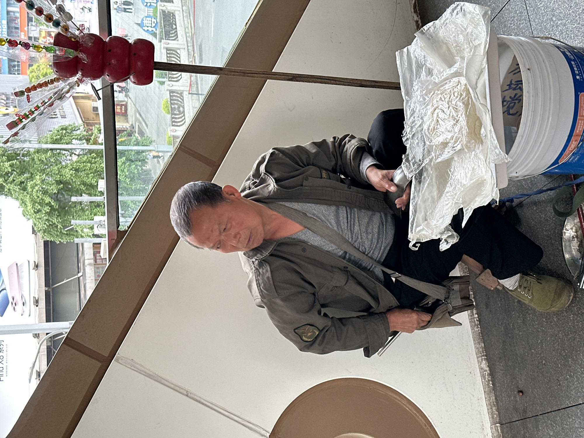 An elderly Asian man sits on a low stool on a city sidewalk in the Jiulongpo District, China. He wears an olive green jacket over a gray shirt, black pants, and laced green canvas shoes. He looks down, manipulating an unidentified white material with his hands. To his right sits a white bucket labeled with Chinese characters and covered with plastic bags. A skewer of candied hawthorns stands to his left. In the background, buildings and trees are visible across the street. A large advertisement is visible to the far left. A round, beige object is cut off at the bottom of the frame.