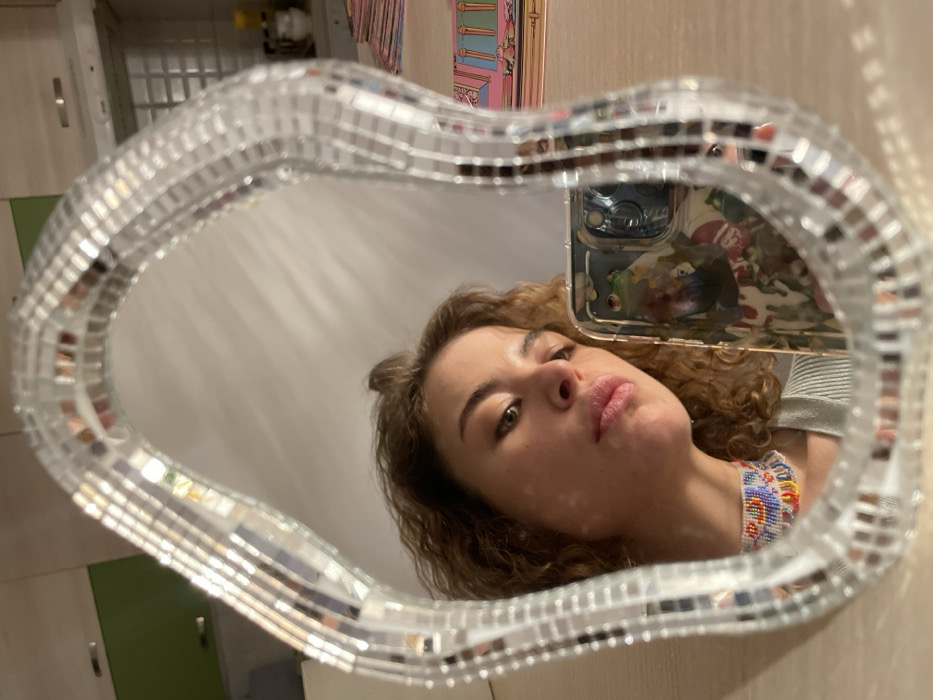 The image depicts the reflection of a female individual taking a selfie in a decorative mirror, observed in an indoor setting in Boreti, Montenegro, consistent with a residential kitchen or dining area.
The central subject is a mirror with an irregular, organic shape, framed by a mosaic of small, rectangular reflective pieces. Reflected within the mirror is the upper body of a woman. She has wavy, light brown hair, and appears to be in her 20s or 30s. Her head is tilted slightly downwards and to the right, with her gaze directed towards the lower left. Her lips are slightly parted, and her expression is neutral. Around her neck is a wide, intricately beaded choker necklace featuring colorful patterns in red, yellow, blue, and white tones. She is wearing a light-colored, possibly ribbed, top.
The woman is holding a smartphone in her right hand (her left hand, as seen in the reflection), which is partially visible in the lower right quadrant of the mirror. The phone has a clear case adorned with a multicolored, abstract design, and a prominent green frog illustration is visible on the case.
The background, visible in the reflection behind the woman, indicates a domestic interior. To the left, a light green wall-mounted cabinet is discernible. To the right, lighter-colored kitchen cabinetry, possibly including an exhaust hood, is visible against a wall. On a surface behind the mirror, a stack of colorful rectangular items, possibly books or cards with illustrations, is present. The overall lighting suggests ambient indoor illumination.