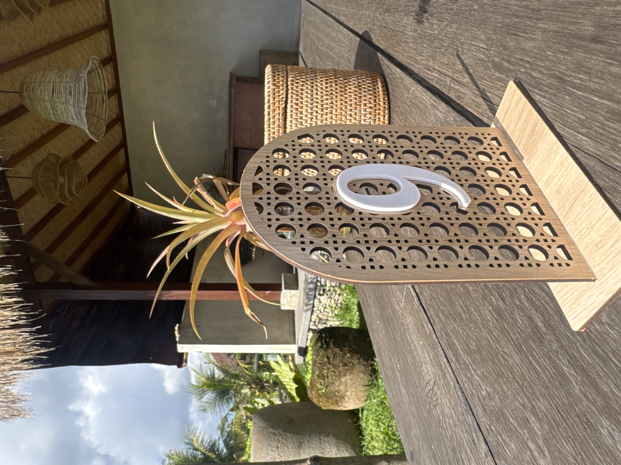 In Tegallalang, Indonesia, a close-up, low-angle shot captures a wooden table number "9" placed on a rustic wooden table. The table number features an arched top with a decorative woven pattern and a white number "9" affixed to its front. In the background, a thatched roof structure with visible wooden beams and woven lampshades is visible. Beyond the structure, lush green foliage and palm trees suggest an outdoor, tropical setting. A woven basket sits to the right of the table number, and two large, weathered stone objects are present in the garden area behind the table. The lighting indicates a bright, sunny day with clear skies and some scattered clouds.