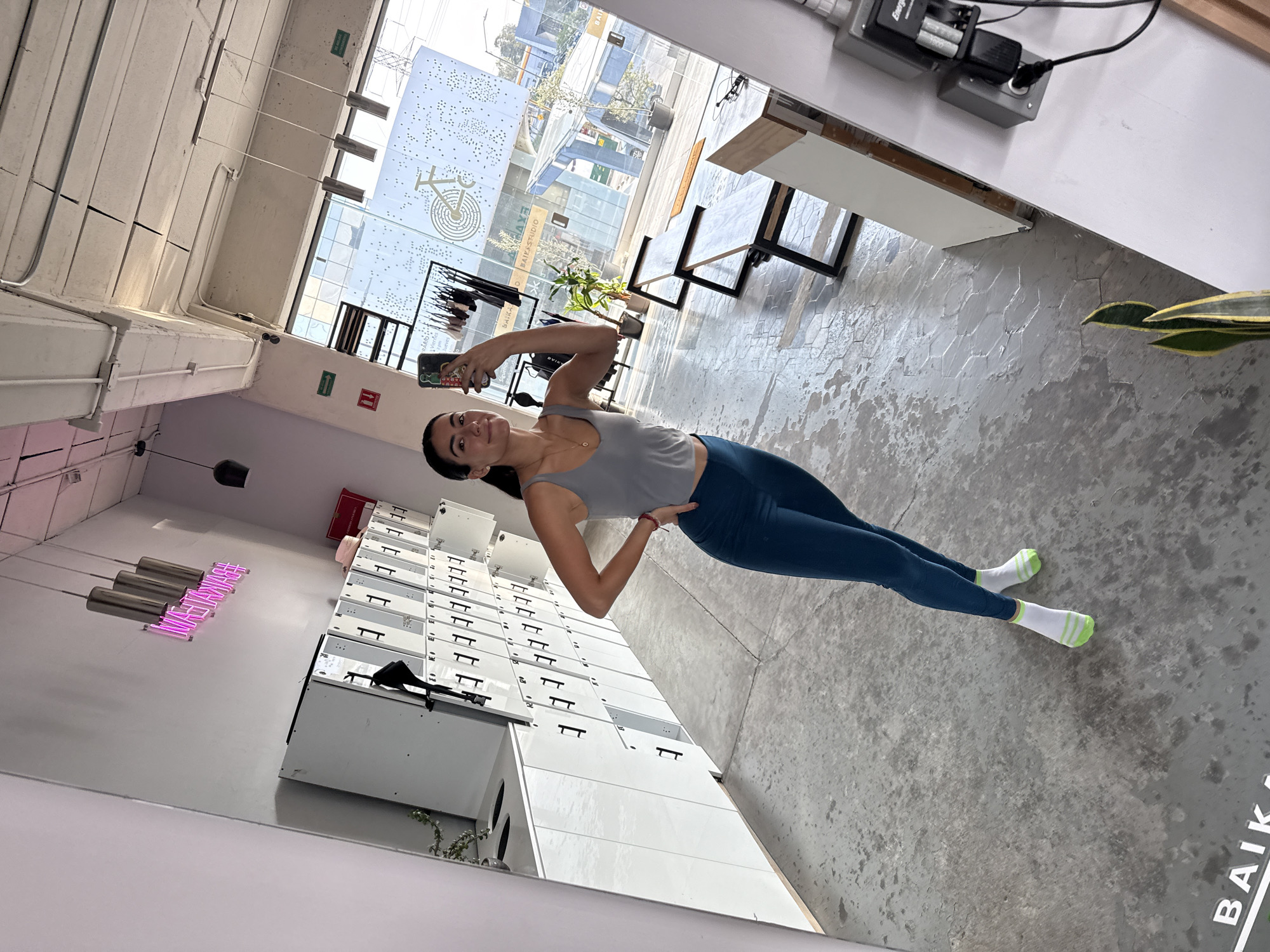 A medium-shot photograph captures a woman taking a selfie inside what appears to be a fitness studio or gym. The location is identified by the logo "BAIKA" reflected in a mirror as being in Puebla, Mexico. The woman is facing the camera, holding her phone up with her left hand to capture the image. She is wearing a gray tank top and blue leggings, with white socks that have a green stripe on the ankle.
The environment includes several features. A wall of lockers is visible behind the woman, and a neon pink sign reading "#baikastudio" is also reflected in the mirror. In the background, there is a large window that looks out onto what seems to be a street scene, featuring other buildings and greenery. The floor has a gray, mottled appearance, possibly concrete or tile. In the far background, clothes are hanging on a rack.