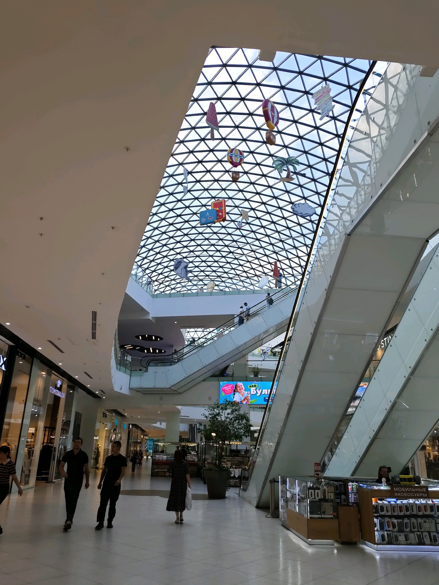 The image shows a modern, multi-level shopping mall interior with a distinctive geodesic dome skylight. The skylight is a prominent architectural feature, composed of triangular glass panels that form a geodesic pattern, allowing natural light to illuminate the space. Suspended from the skylight are decorative elements resembling hot air balloons and other whimsical shapes, adding a playful touch.
Inside the mall, there are escalators and walkways connecting different levels, with people visible on them. Several storefronts line the ground floor, one of which displays the "TOM TAILOR" brand. A mobile accessories kiosk is also visible, stocked with numerous phone cases and related items. A large digital billboard is mounted on a wall, displaying colorful advertisements.
The flooring appears to be a polished tile, reflecting the ambient light. The overall atmosphere suggests a busy and contemporary commercial environment. The presence of a billboard with Russian text, specifically "НАШ БУЛЬВАР" (Nash Bulvar, meaning "Our Boulevard") and other Cyrillic lettering, strongly indicates the location is within Russia. Given that "Bulvar" is a common element in Russian shopping mall names, and without further context to differentiate it from other Russian locations, the mall is identified as "Nash Bulvar" in Rostov-on-Don, Russia.