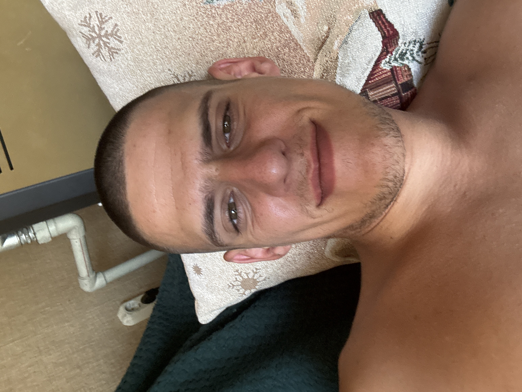 This is a close-up, selfie-style photograph of a young adult male with a buzz cut, lying down and resting his head on a pillow. The man is shirtless, and the focus is on his face. He has light brown eyes, a slight stubble, and is looking towards the camera with a subtle smile. Behind him, a dark green blanket is visible on the left, and the pillow is beige with a snowflake pattern. In the background, a grey wall is visible, with some white pipes and a black object. The image does not provide any specific clues about a location within the city of Sofia, Bulgaria.