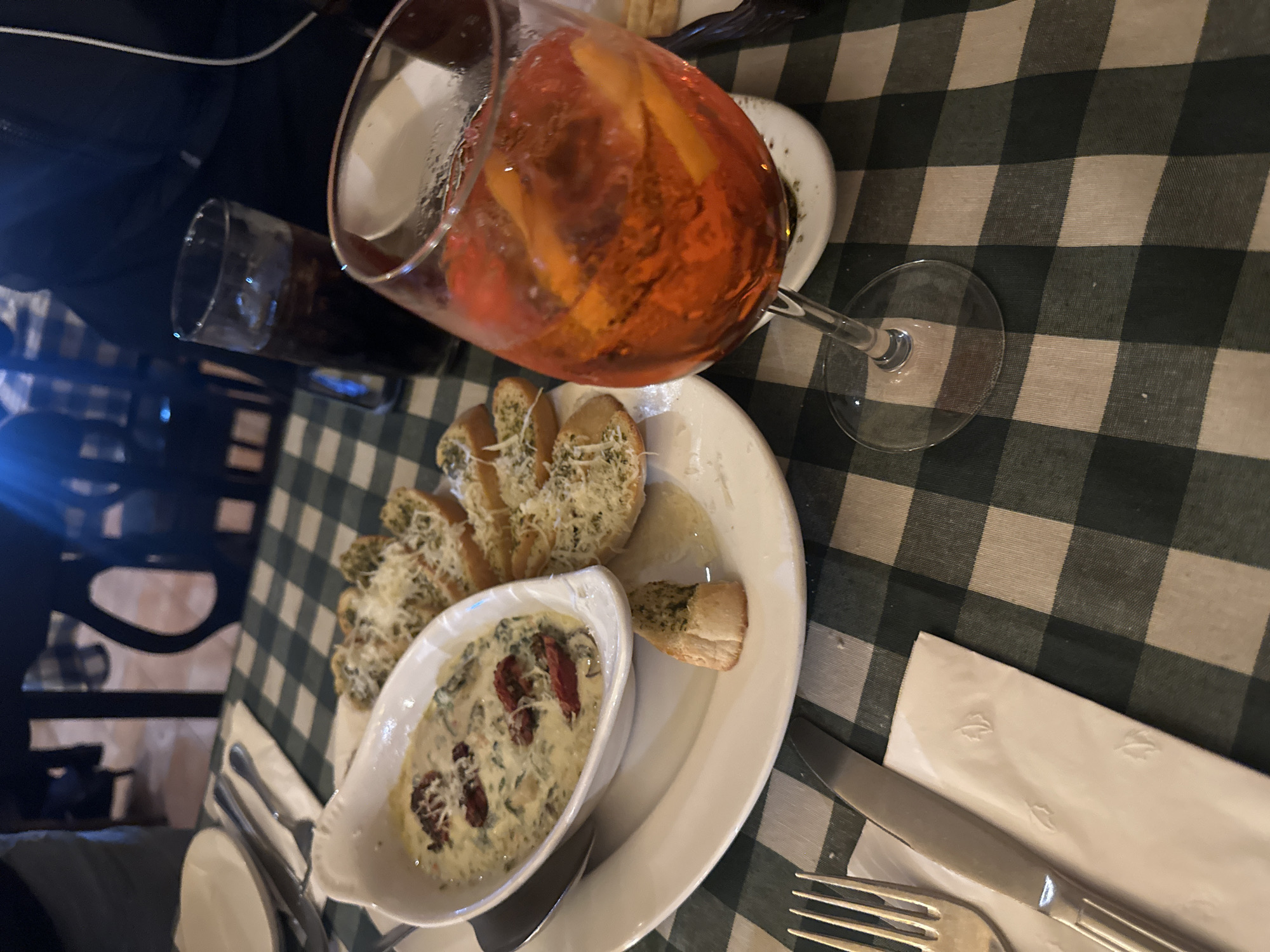 The image depicts a table setting in what appears to be a restaurant in Naucalpan de Juárez, Mexico. A green and white checkered tablecloth covers the table. Dominating the scene is a large glass of an orange-colored beverage with what appears to be orange slices inside, indicating an Aperol Spritz. Adjacent to the glass, there is a white plate containing a bowl of spinach and artichoke dip garnished with sun-dried tomatoes. Arranged around the dip are slices of garlic bread, some topped with cheese or herbs. Cutlery consisting of a fork and knife are laid out on a white napkin. A dark-colored beverage in a glass is also visible. The scene is indoors.