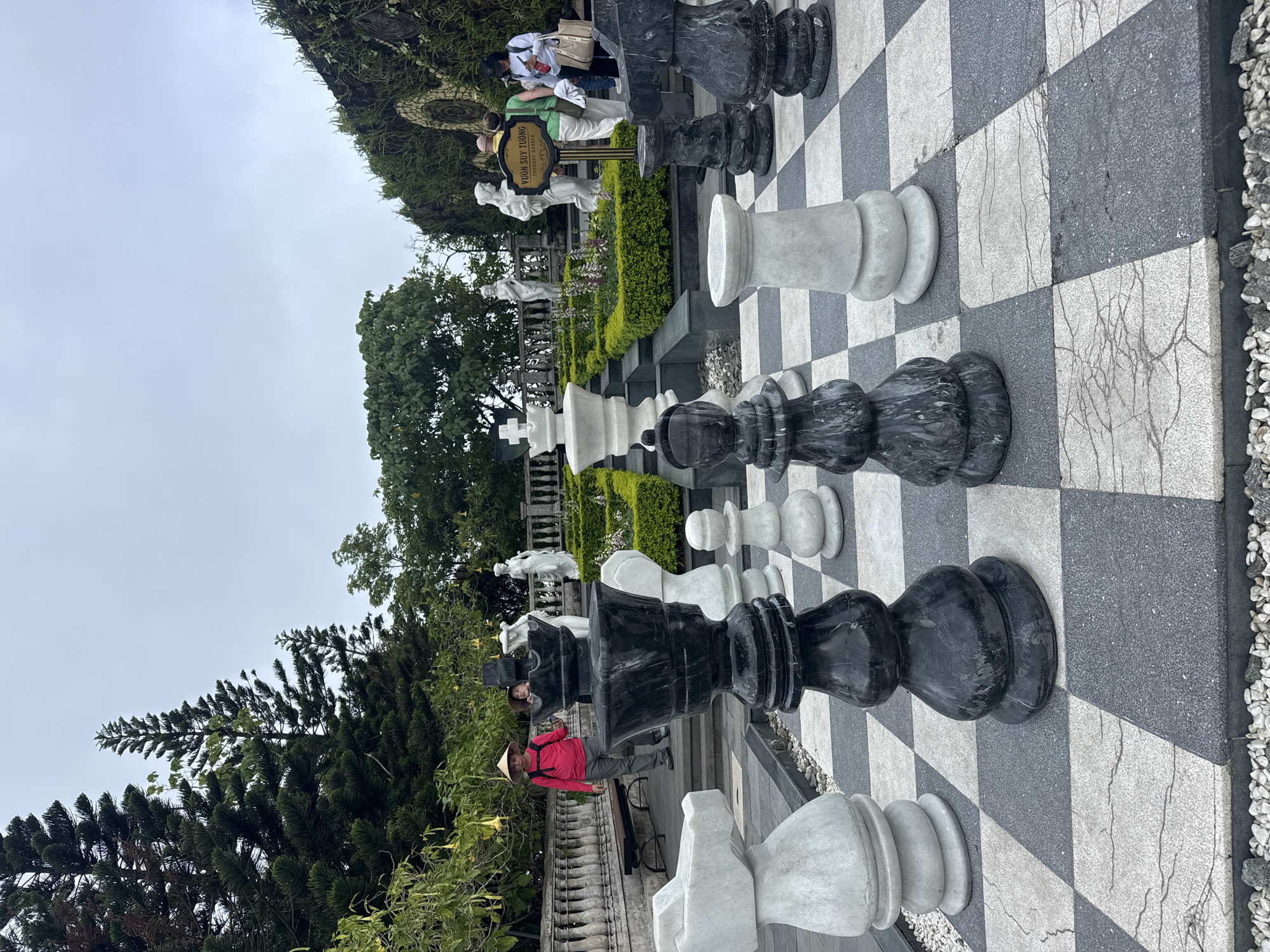 The image shows a chessboard with oversized chess pieces in the park, located in the city of Đà Nẵng, Vietnam. The black and white chess pieces are arranged on a black and white checkered stone surface. A man wearing a red shirt and a conical Asian hat stands next to the chessboard. In the background, there are trees, bushes, and statues. Several other people are standing behind a sign. The sky is overcast.